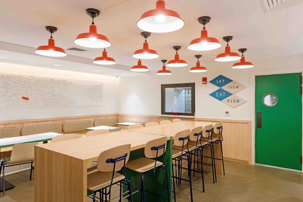 Modern dining area with wooden tables and chairs, red ceiling lights, green door, and wall art saying "Let them eat fish."