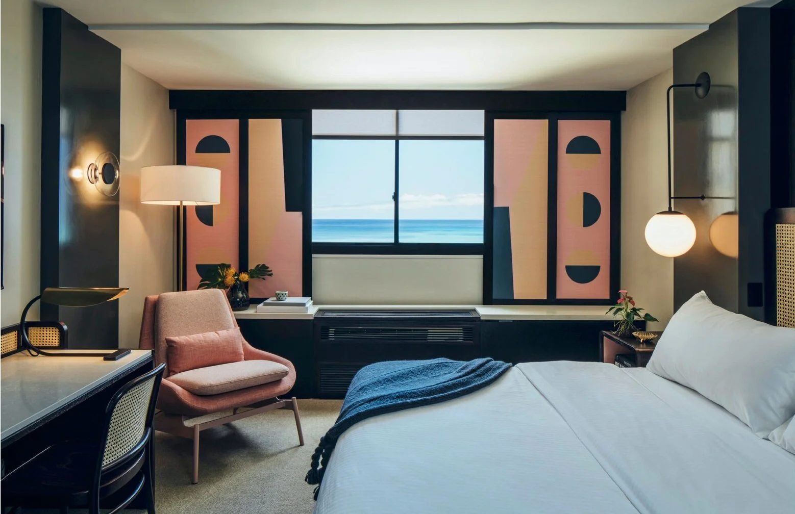 Modern hotel room interior with a bed, armchair, desk, and ocean view window. Minimalist decor with geometric art, ambient lighting, and blue blanket on bed.
