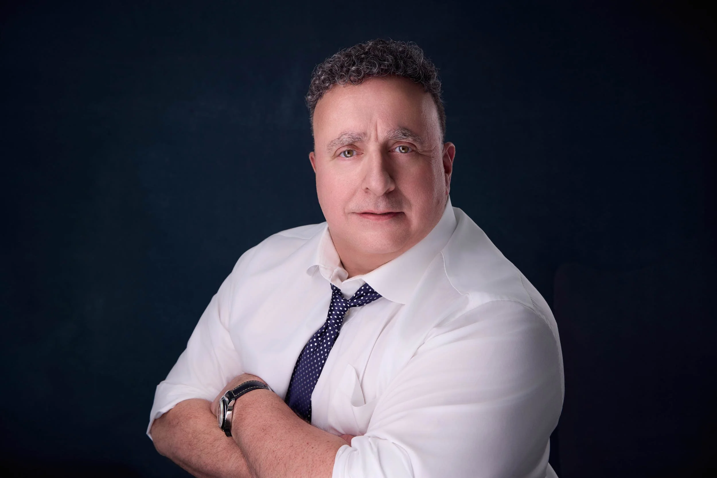 Professional portrait of a man with rolled sleeves and folded arms against a deep blue background.