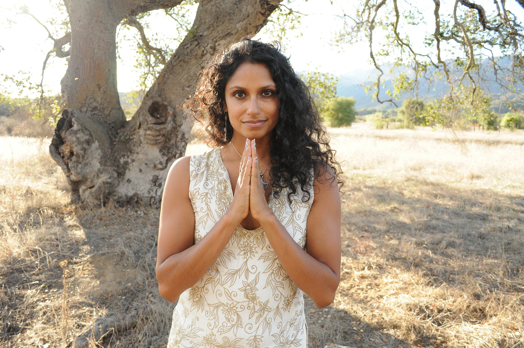 Woman standing outdoors near a tree with her hands pressed together in a prayer pose, wearing a white dress with gold floral patterns, surrounded by a natural landscape with grass and distant trees.