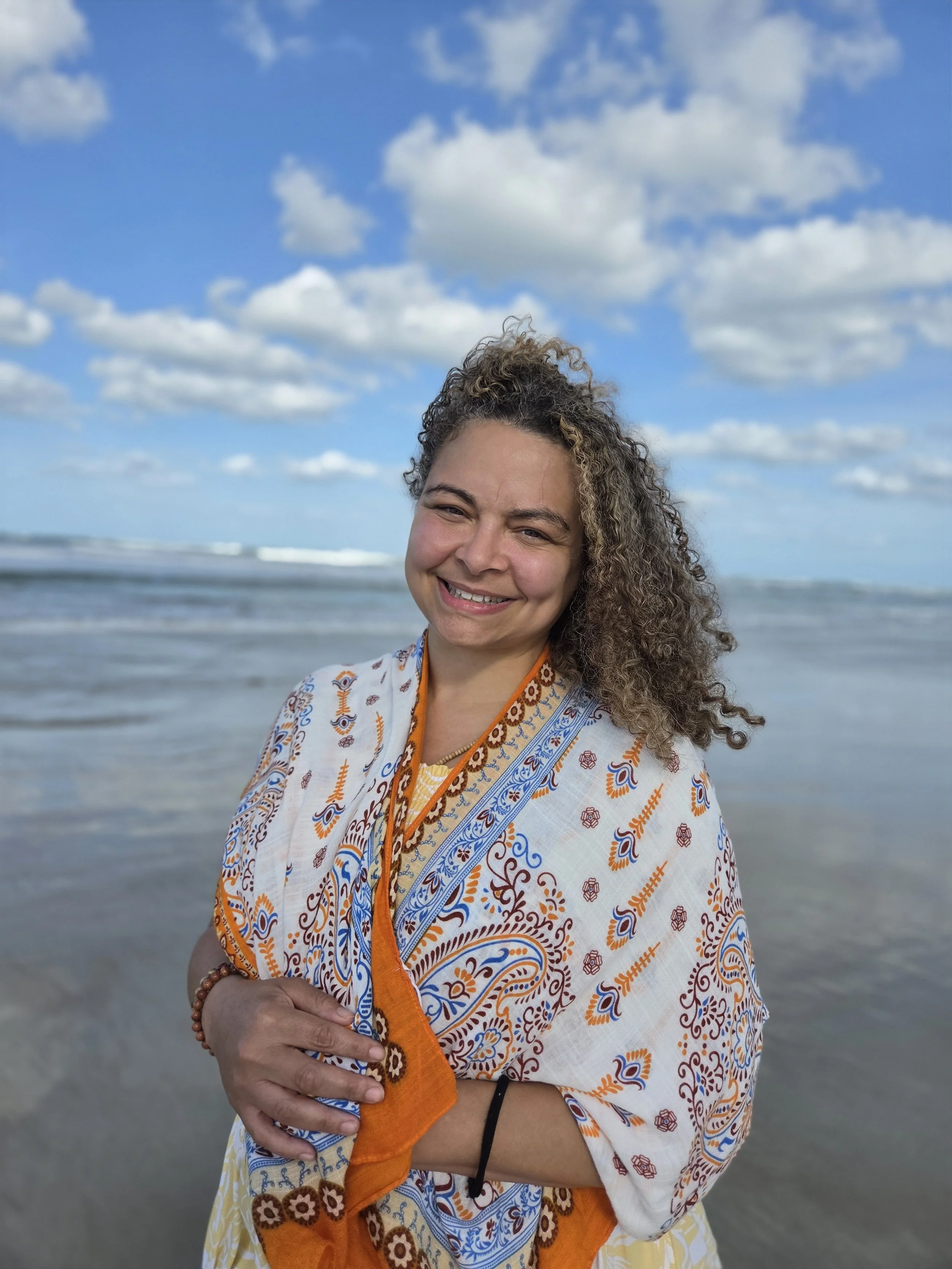 Young woman with curly hair smiling and extending her arms outdoors against a partly cloudy sky and greenery.