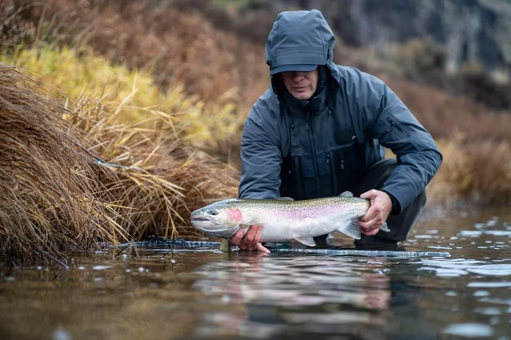 Man in a dark jacket and hooded raincoat holding a large rainbow trout in a shallow stream with brown grass on the banks.
