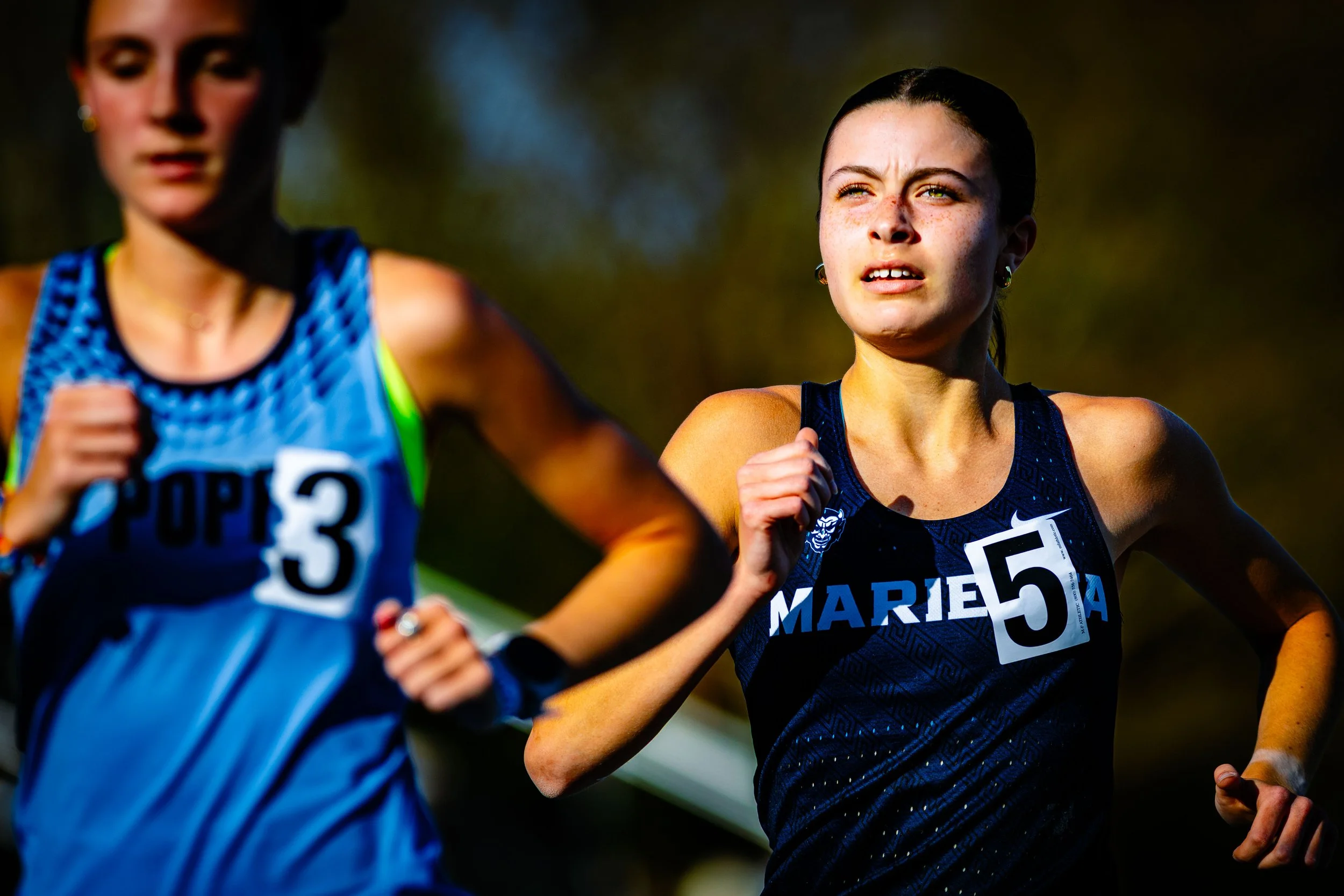 Two female runners competing in a race, wearing athletic gear and race bibs, with one in focus with race number 5 and the other partially visible with race number 3.