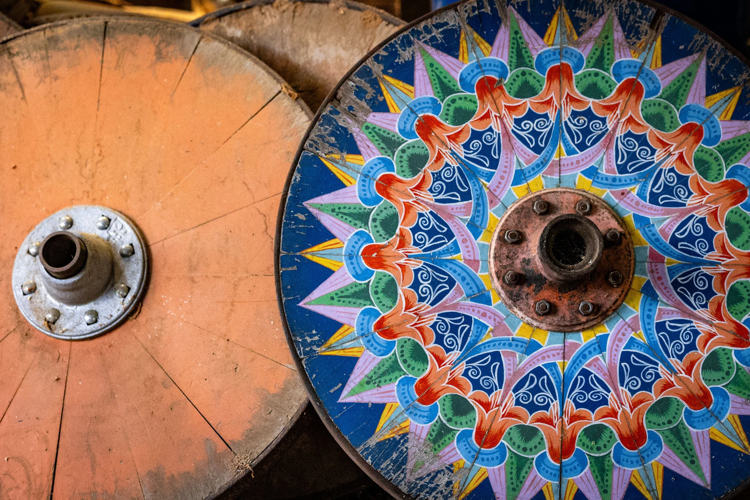 Close-up of two colorful, vintage, spinning-mill wheels with one painted with a vibrant geometric floral pattern, showing signs of wear and rust.