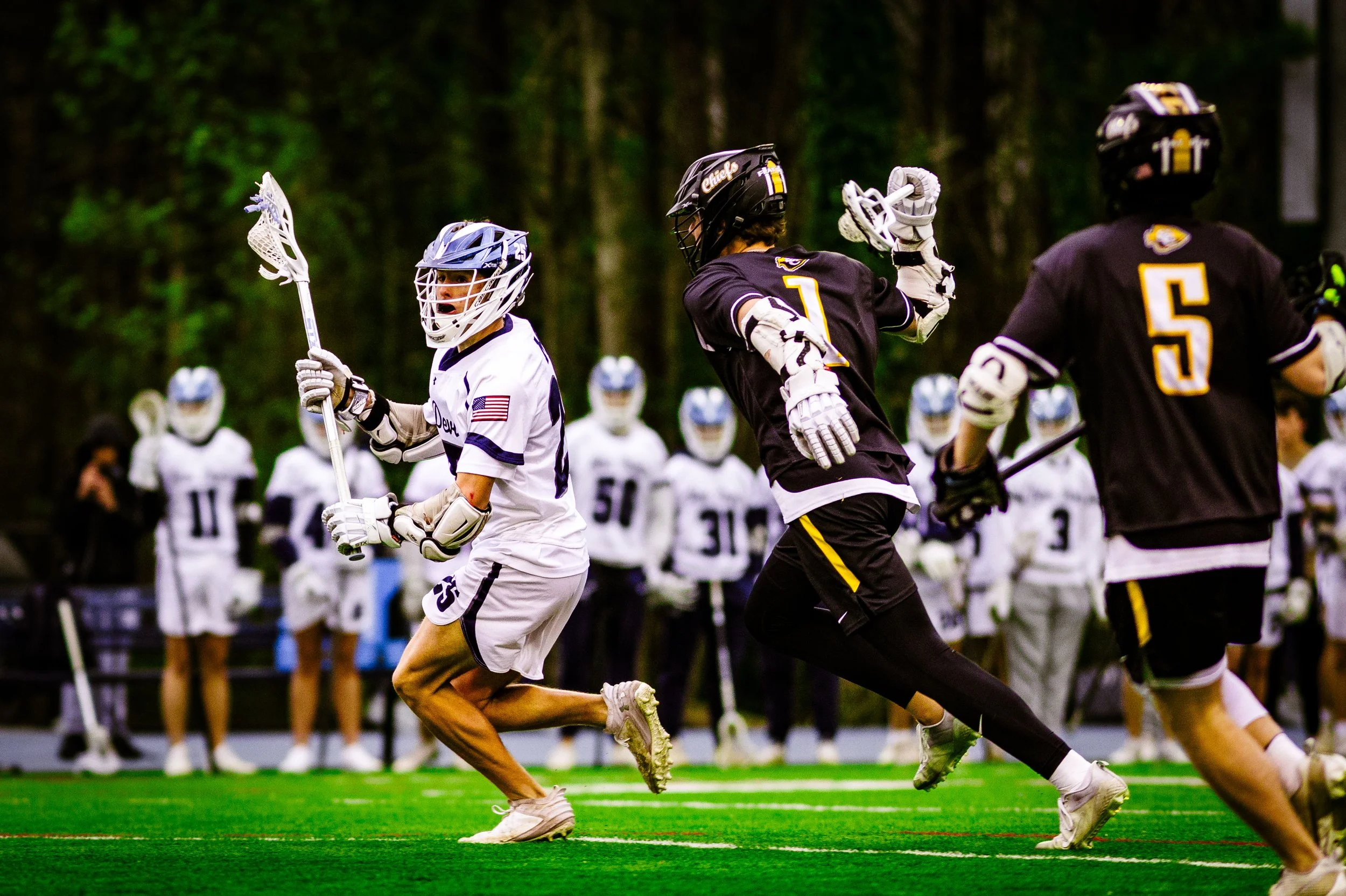 Lacrosse players on field during game, one in white jersey holding lacrosse stick, others in black jerseys running, with team in background and trees around playing area.