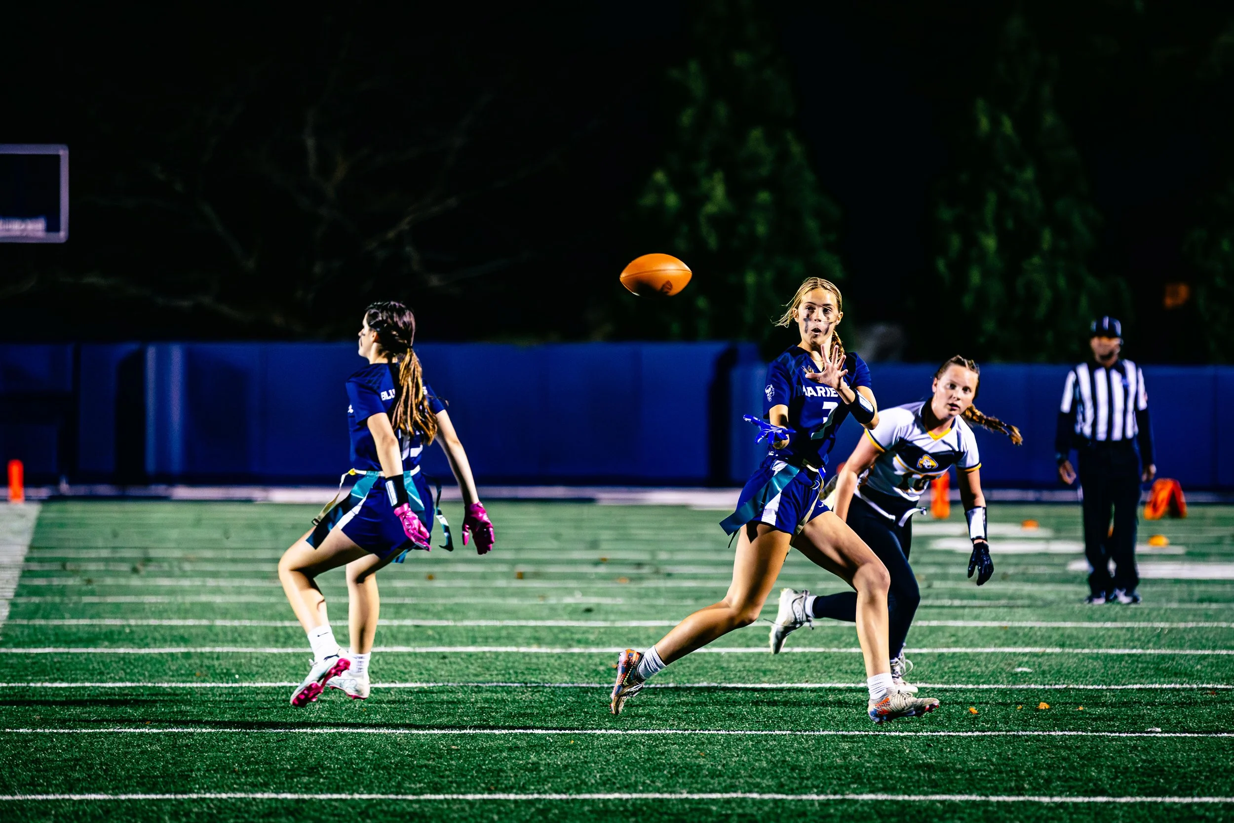Female flag football players on a field at night, one with the ball, others moving to catch or defend, referee in background.