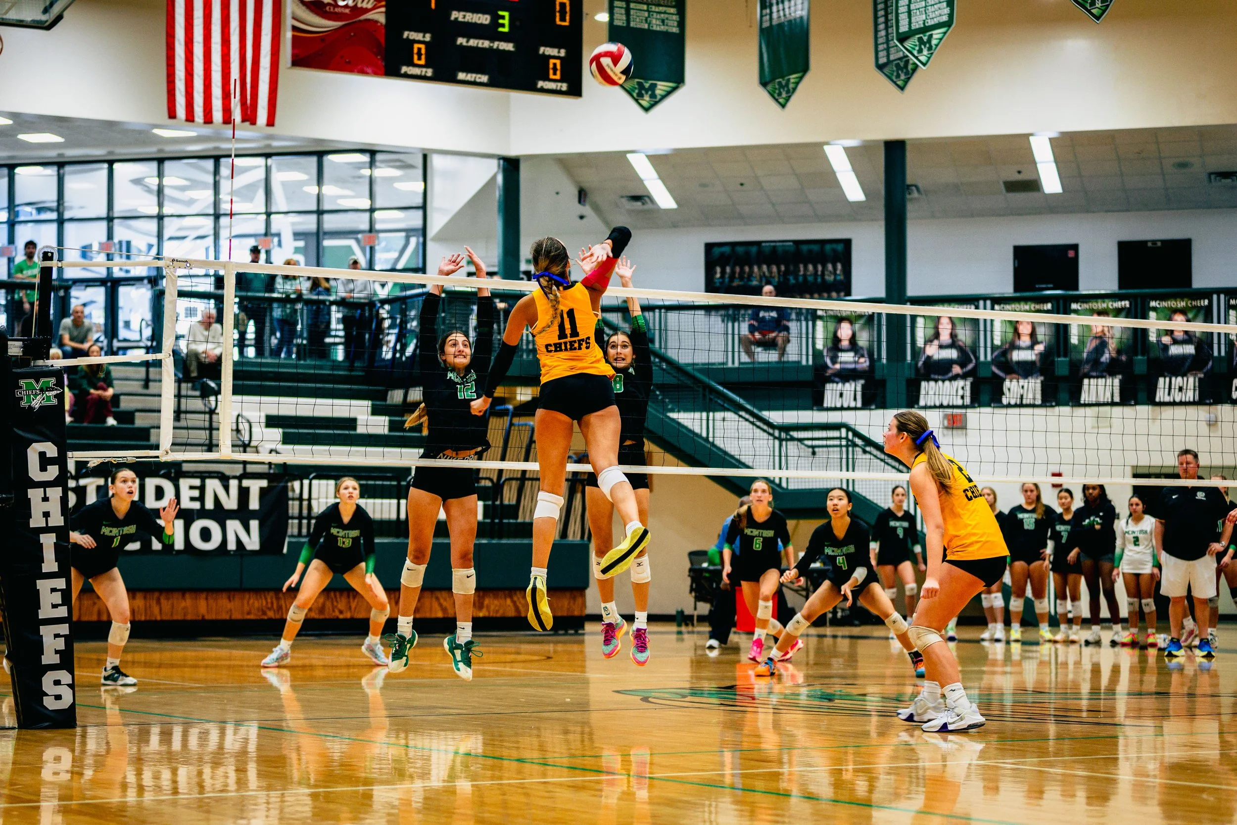 Girls' volleyball game in progress, with players jumping to hit the ball over the net in a gymnasium, with spectators sitting in the background.