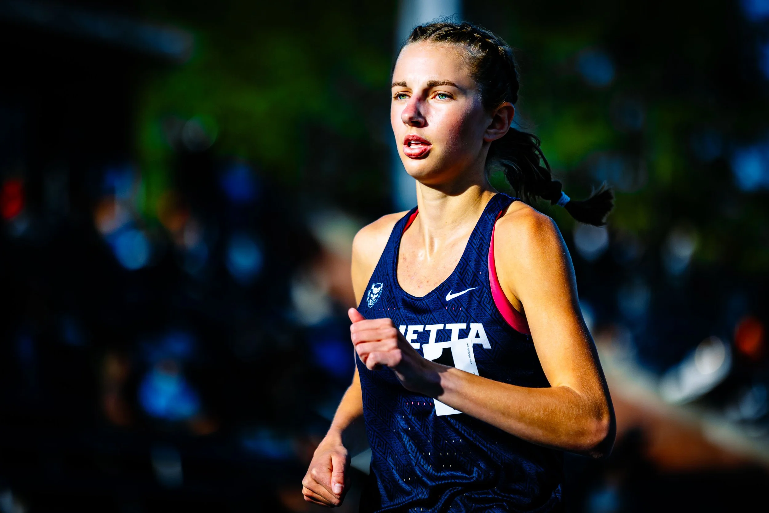 A young woman running in a race, wearing a navy blue athletic tank top with white and pink accents.