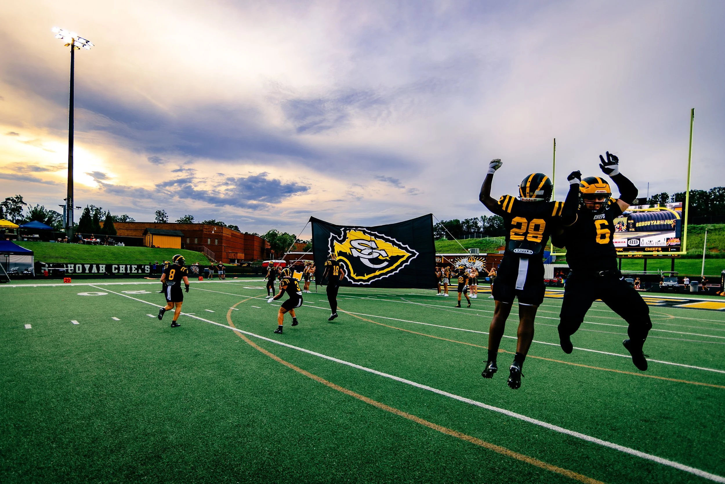 Football players in black and yellow uniforms celebrating on the field, with some jumping and holding a team flag, during a game at sunset.