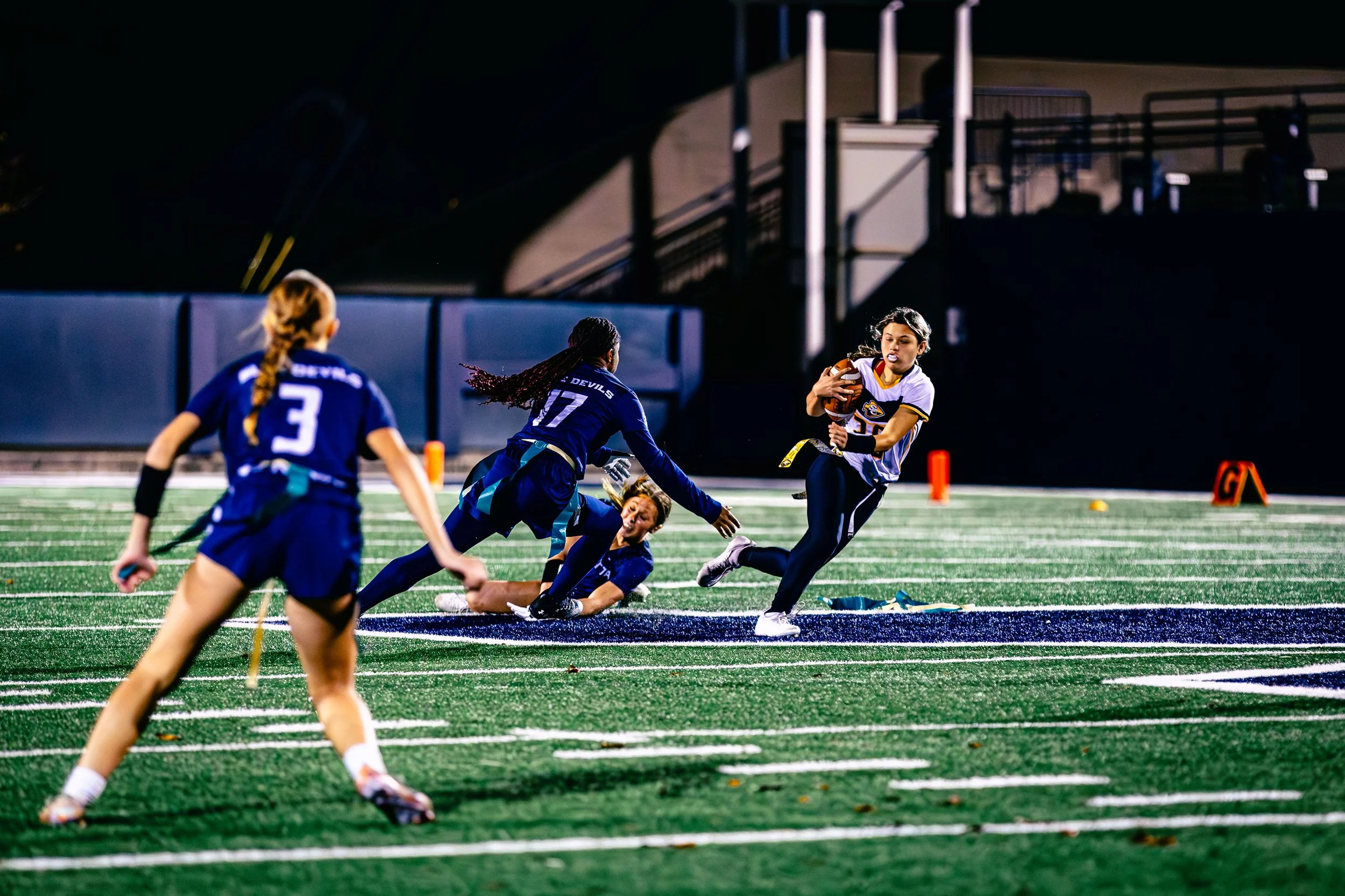 A group of girls playing flag football on a field at night, with one girl holding the football and running while others attempt to tackle her.