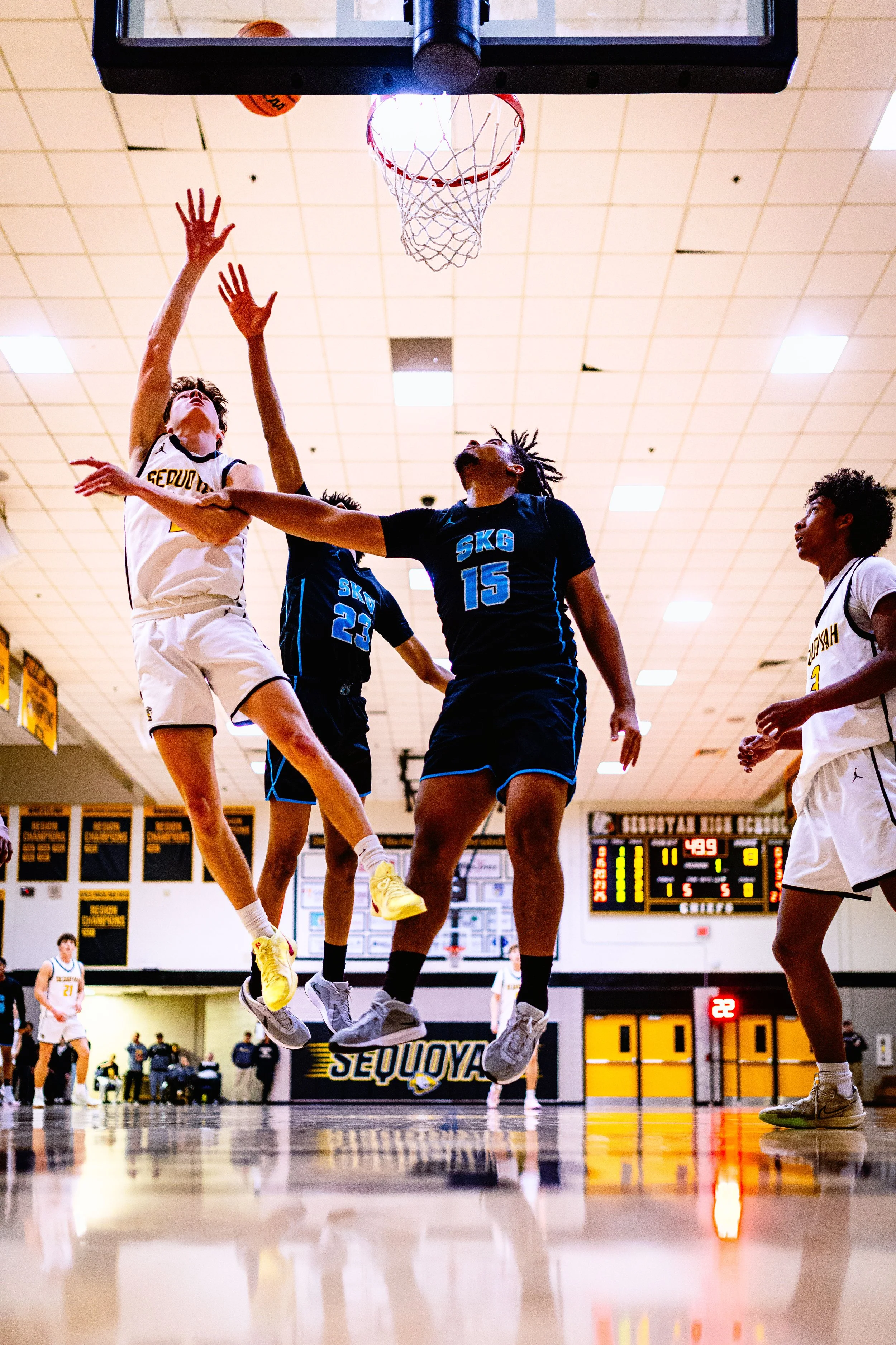A basketball game in progress with players jumping near the hoop, one player in a white jersey and two players in black jerseys trying to block the shot. The game is played in a gym with a scoreboard and banners in the background.