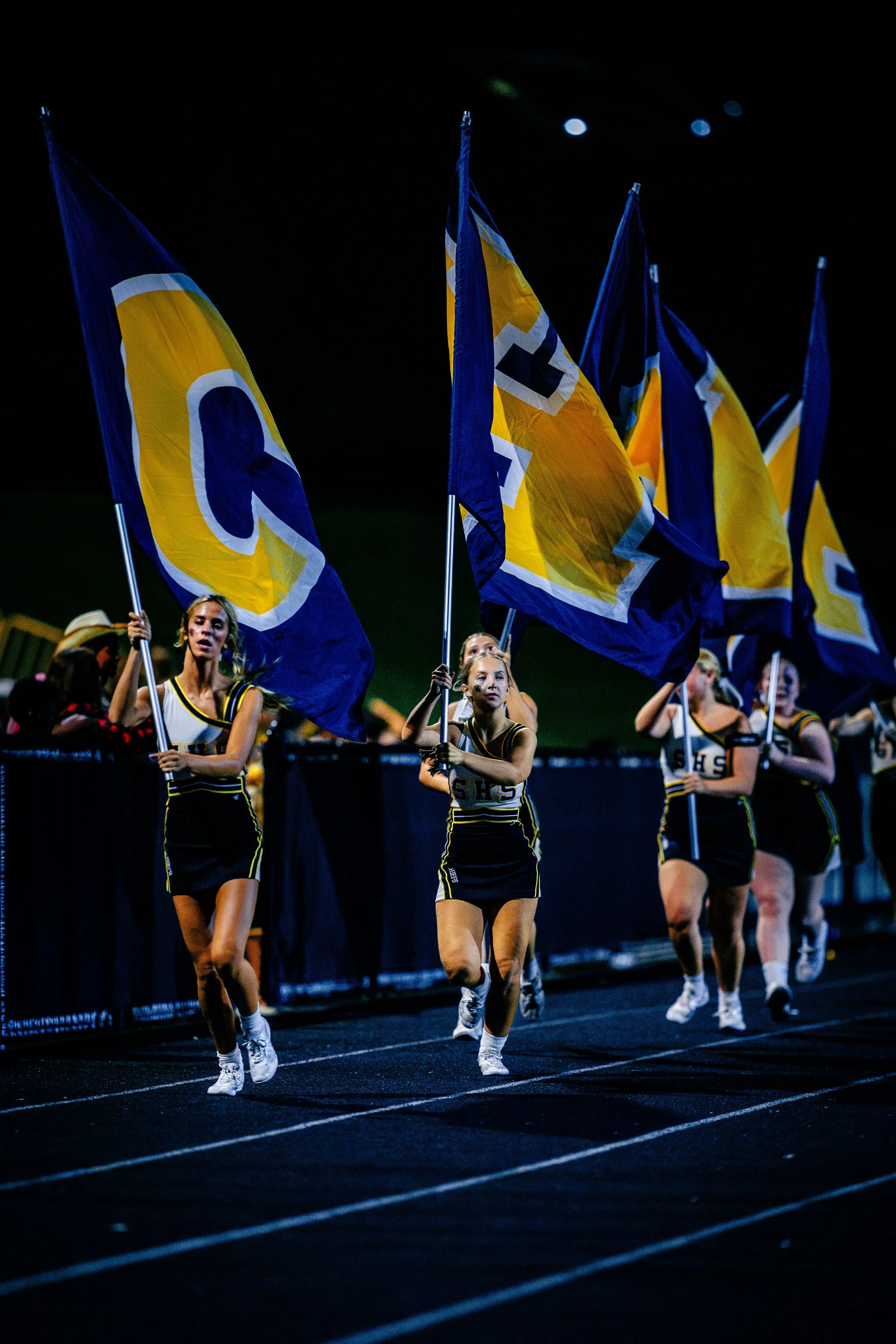 Cheerleaders in black and yellow uniforms holding blue and yellow flags during a nighttime sporting event on a track.