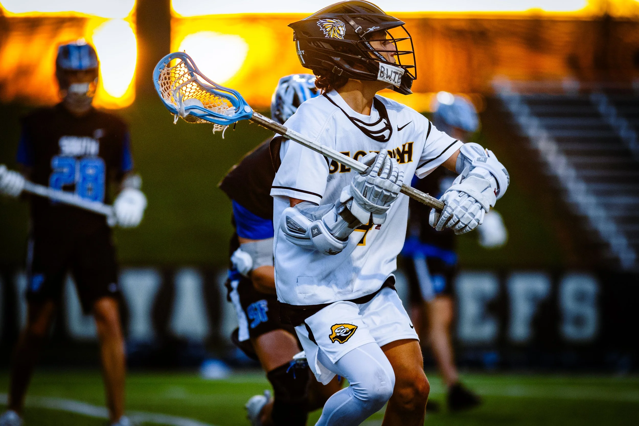 Lacrosse player in white uniform running with a lacrosse stick, wearing a black helmet, with other players in the background during sunset.