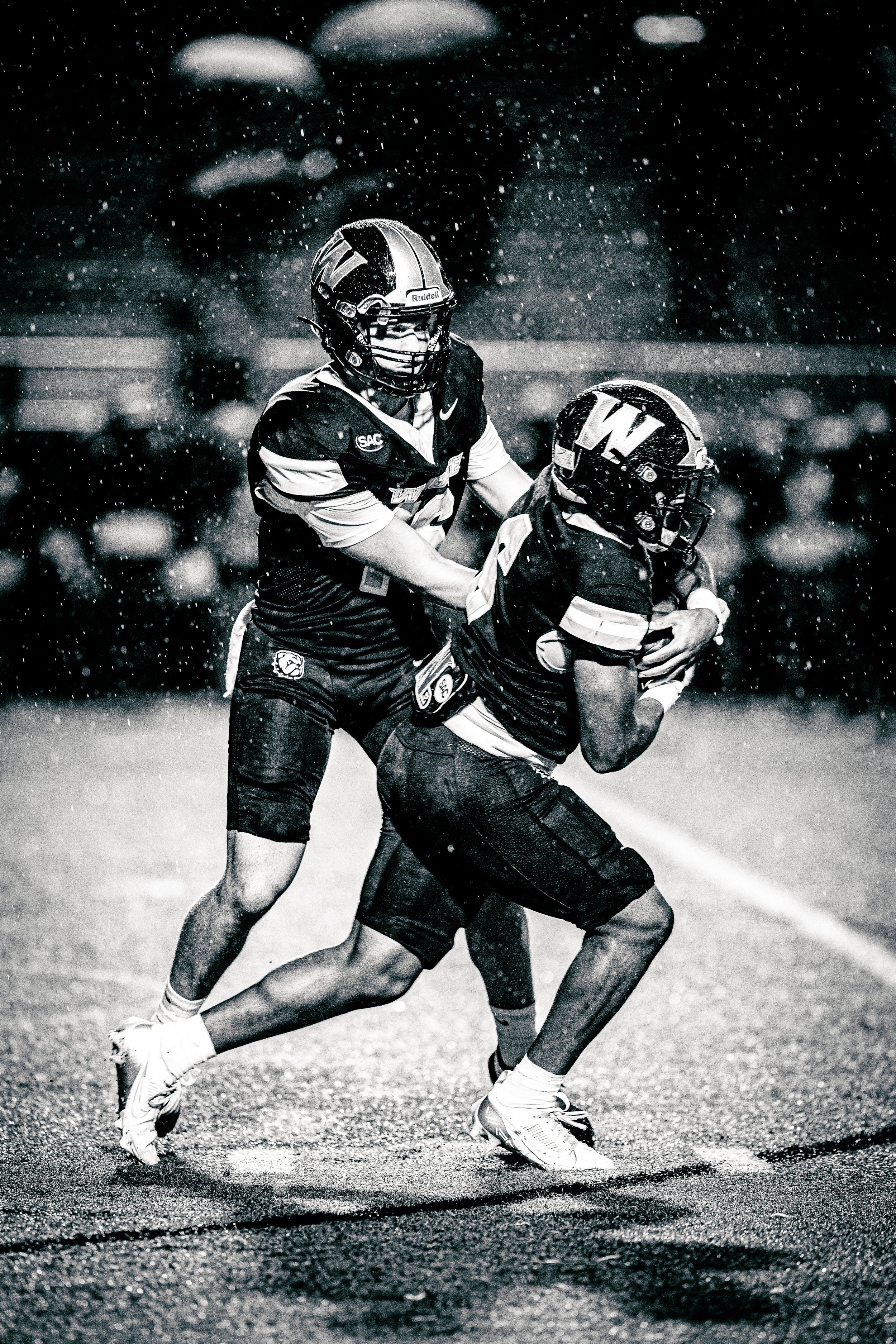 Black and white action shot of two football players in rain, one wearing a helmet with a 'W' logo, tackling the other on the field.