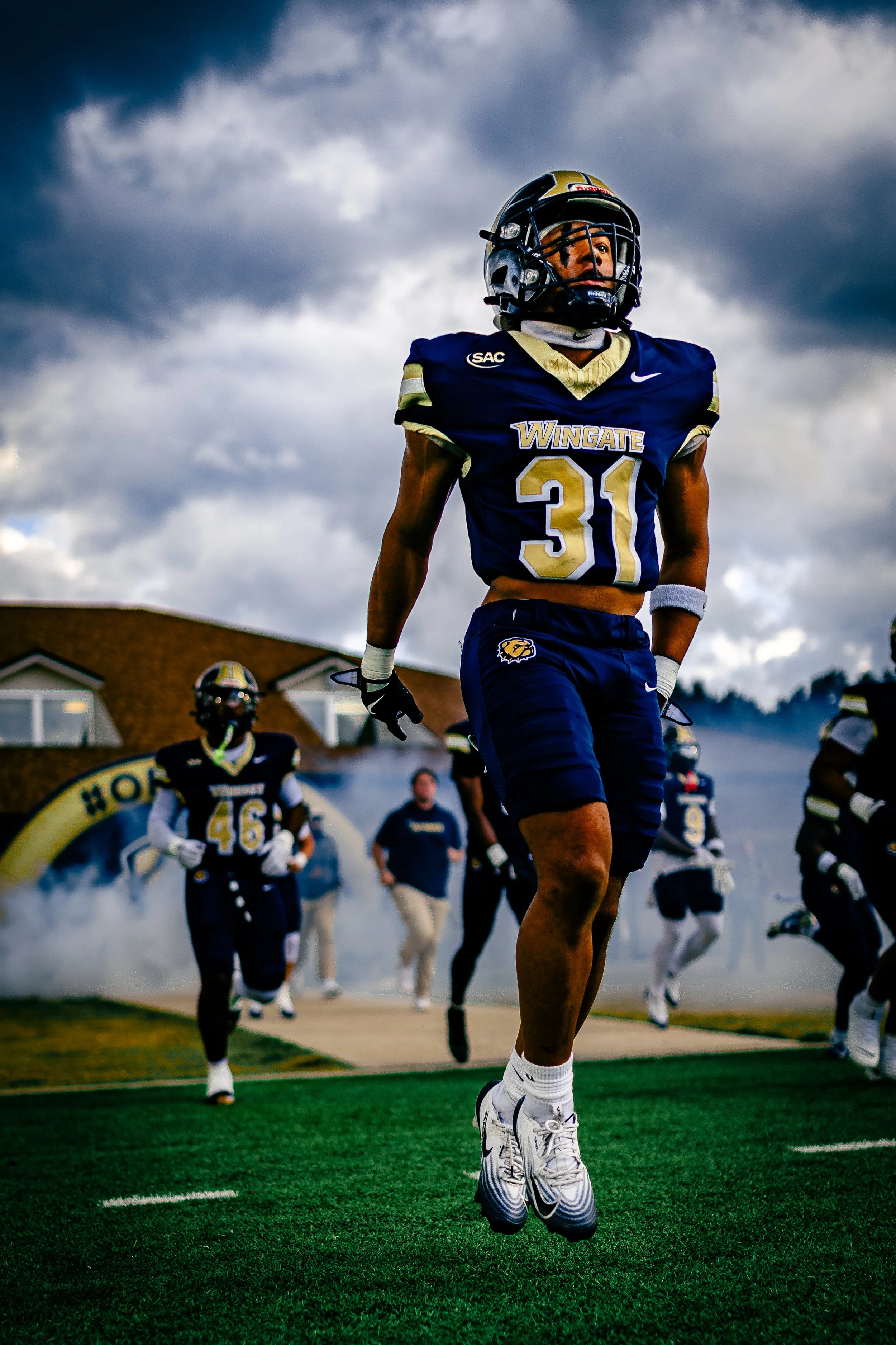 American football player wearing number 31 in a blue and gold team uniform, jumping in the air during a game with teammates and opponents in the background, under a cloudy sky.