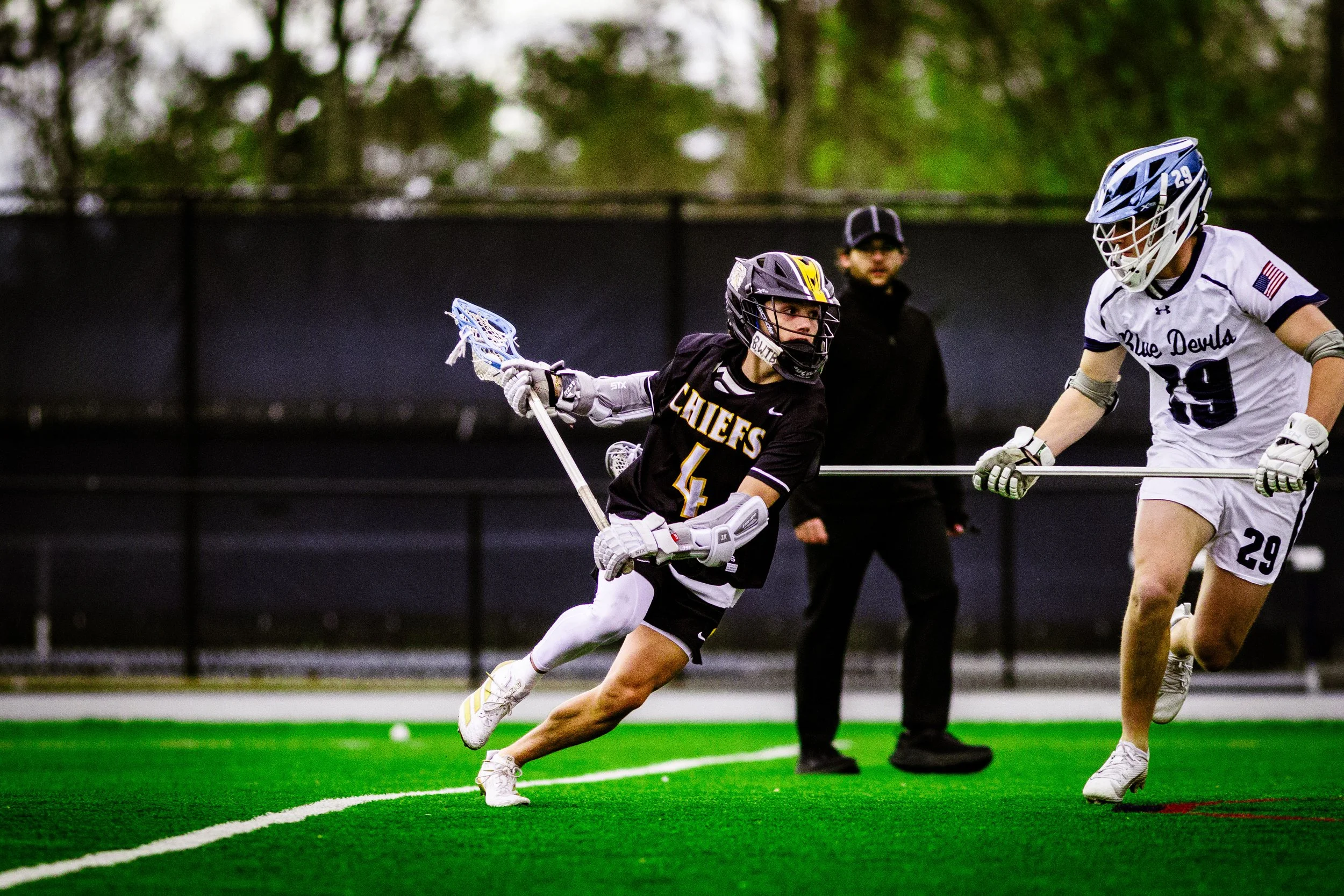 Lacrosse game with two players in action on a green turf field, one in black uniform with 'Chiefs' and number 4, the other in white uniform with 'Blue Devils' and number 29, with a coach or referee watching in the background.
