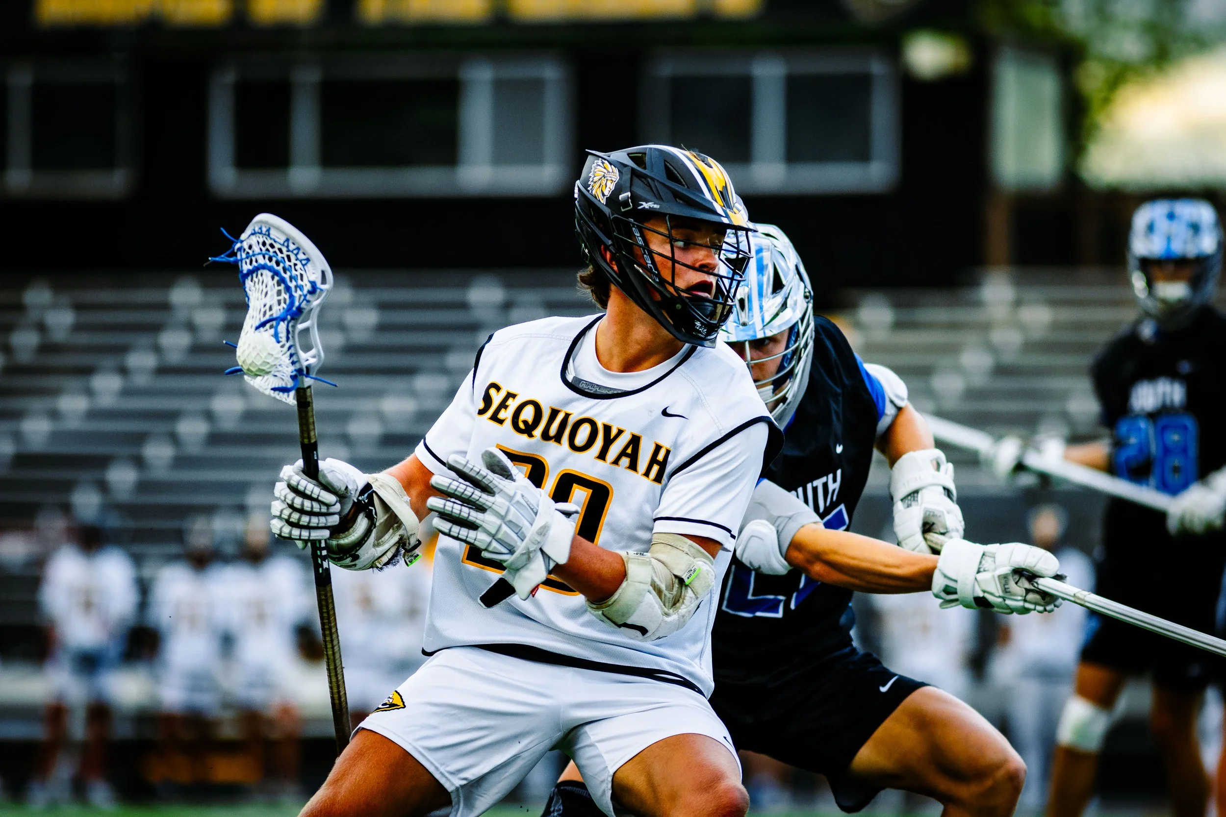 Lacrosse players in action on the field, one from Sequoiah in white jersey and helmet, and another from North Carolina in black jersey, both wearing helmets and gloves, with lacrosse sticks.