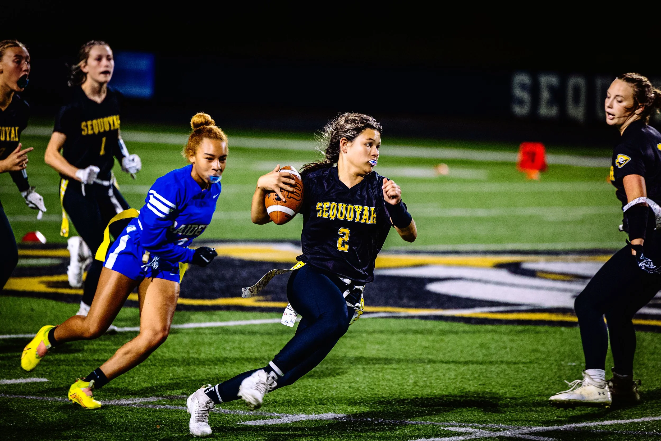 Women playing flag football on a field at night, with one woman holding a football and running, others chasing or defending, with a logo on the field and a scoreboard in the background.