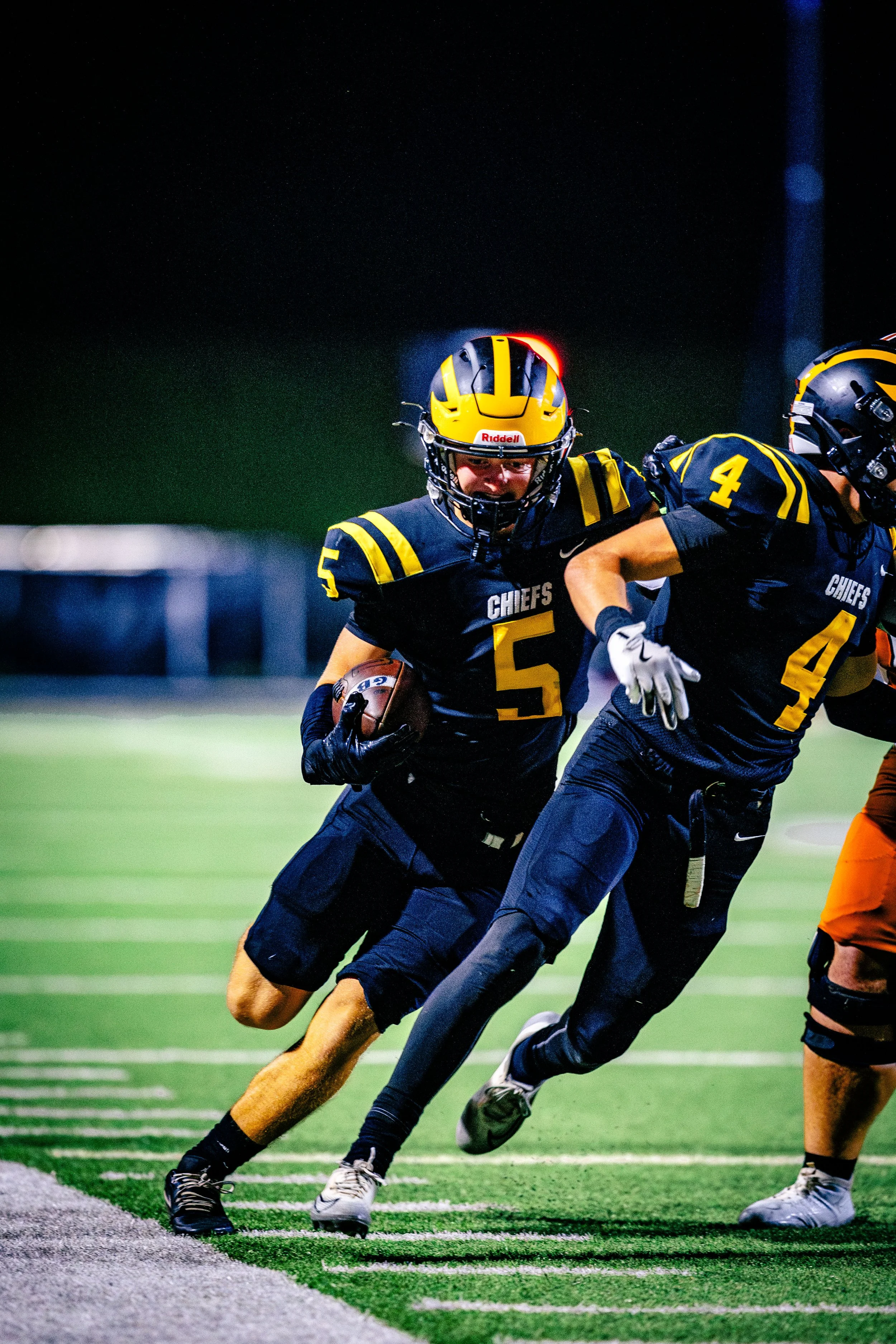 Two football players in black and yellow uniforms from the Chiefs team play during a game at night; one is running with the ball while the other blocks an opponent.