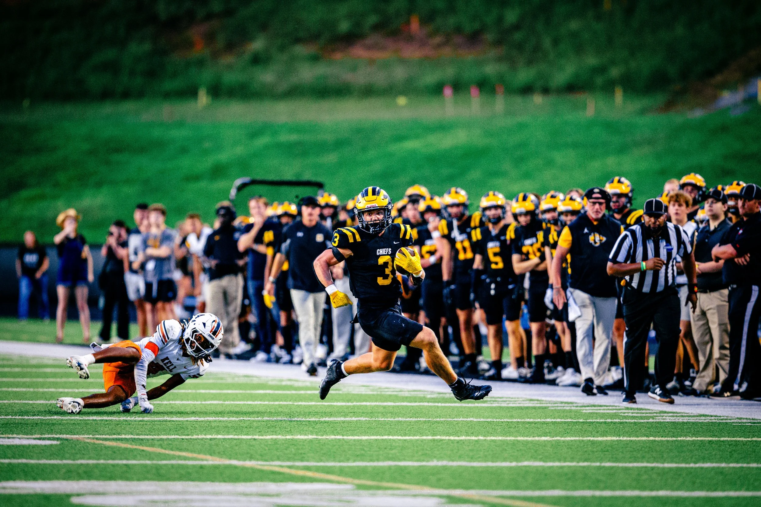 American football game scene with player running with the ball near the sideline and opposing player falling on the ground, with teammates and coaches watching from the sideline.
