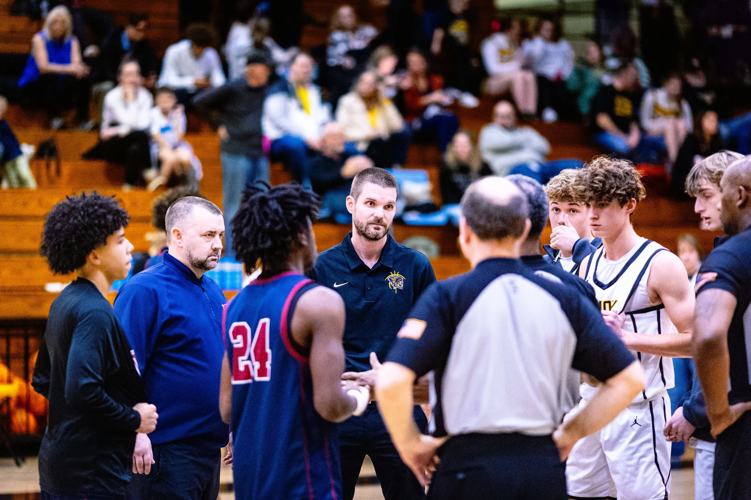 Basketball players and referees gathered in a gymnasium for a game, with spectators in the background.
