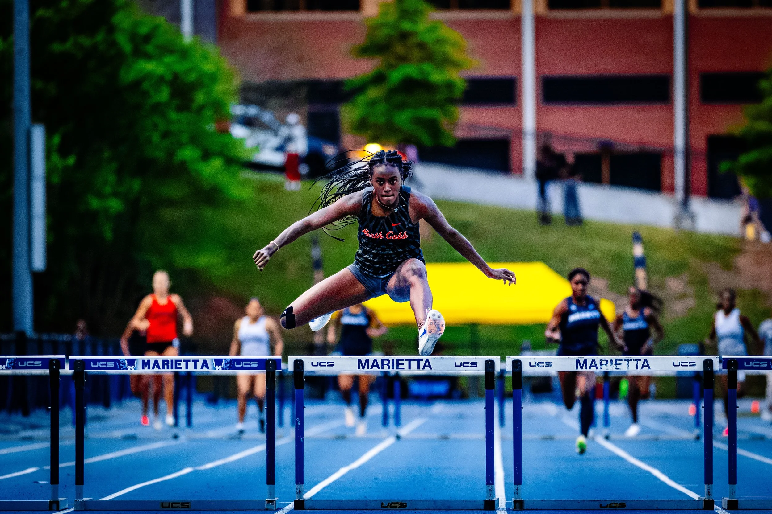 A female hurdler in athletic gear jumping over a hurdle during a race at a track meet. Other competitors are visible in the background.