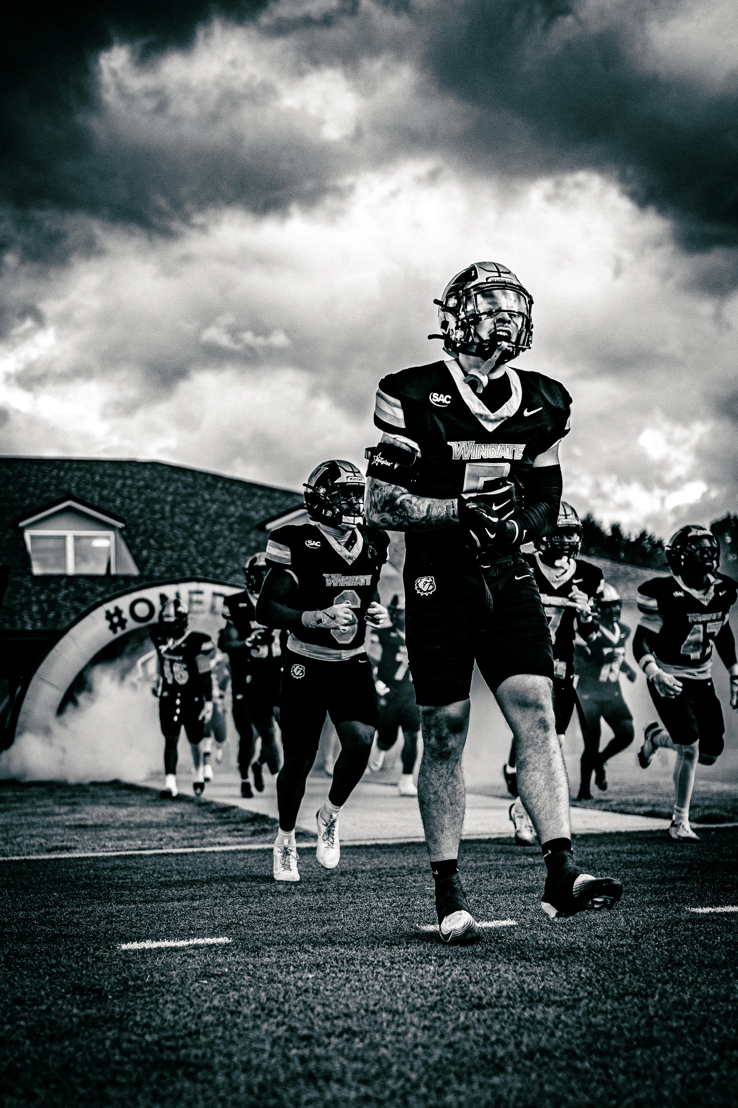 Black and white photo of a football team running under an arched banner that reads '#ONE'. The player at the front is wearing a helmet, gloves, and a jersey with 'WINGATE' and a number, and appears excited or focused. The scene takes place outdoors o