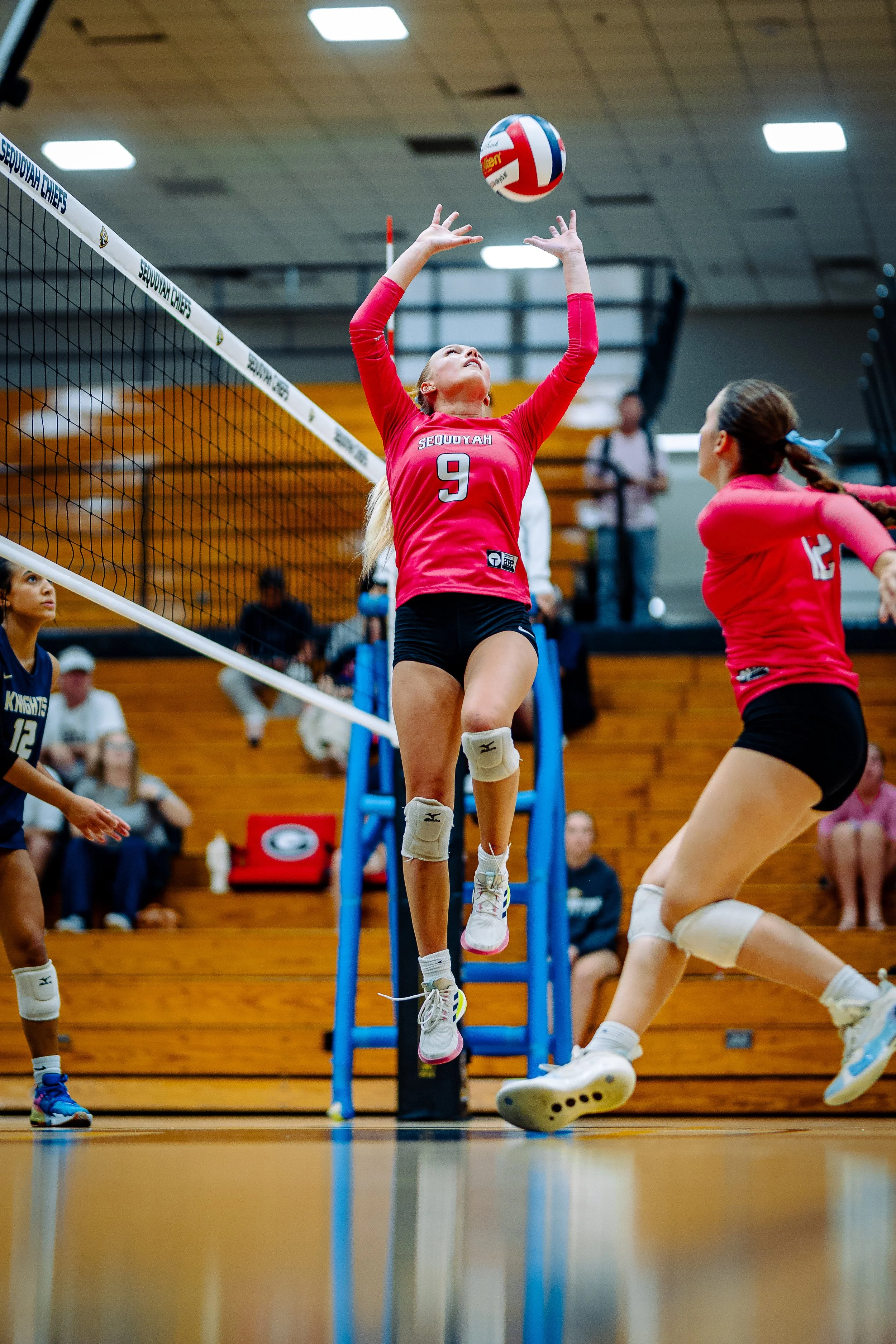 A female volleyball player wearing a pink jersey with the number 9 is jumping to hit the ball at the net, with her arms raised and eyes focused on the ball. Other players and spectators are visible in the background in a gymnasium.