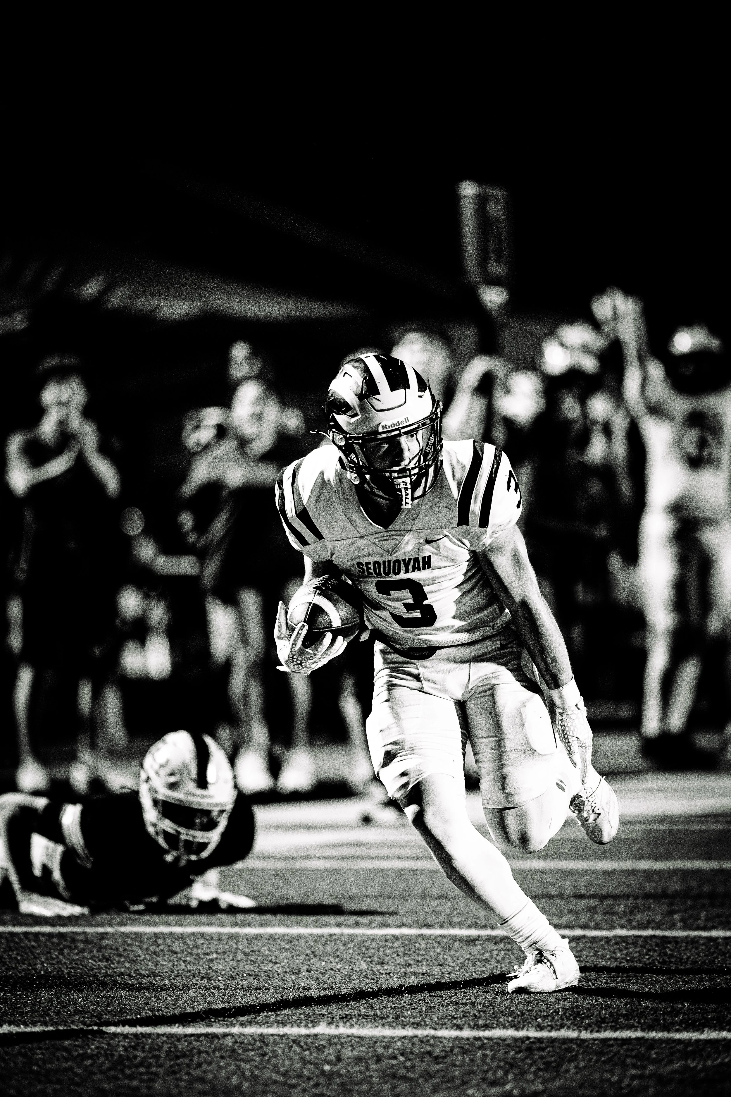 A football player wearing a helmet and jersey with the text 'SEQUOYAH' is running with the ball, while a player from the opposing team is on the ground. Fans and spectators are blurred in the background.