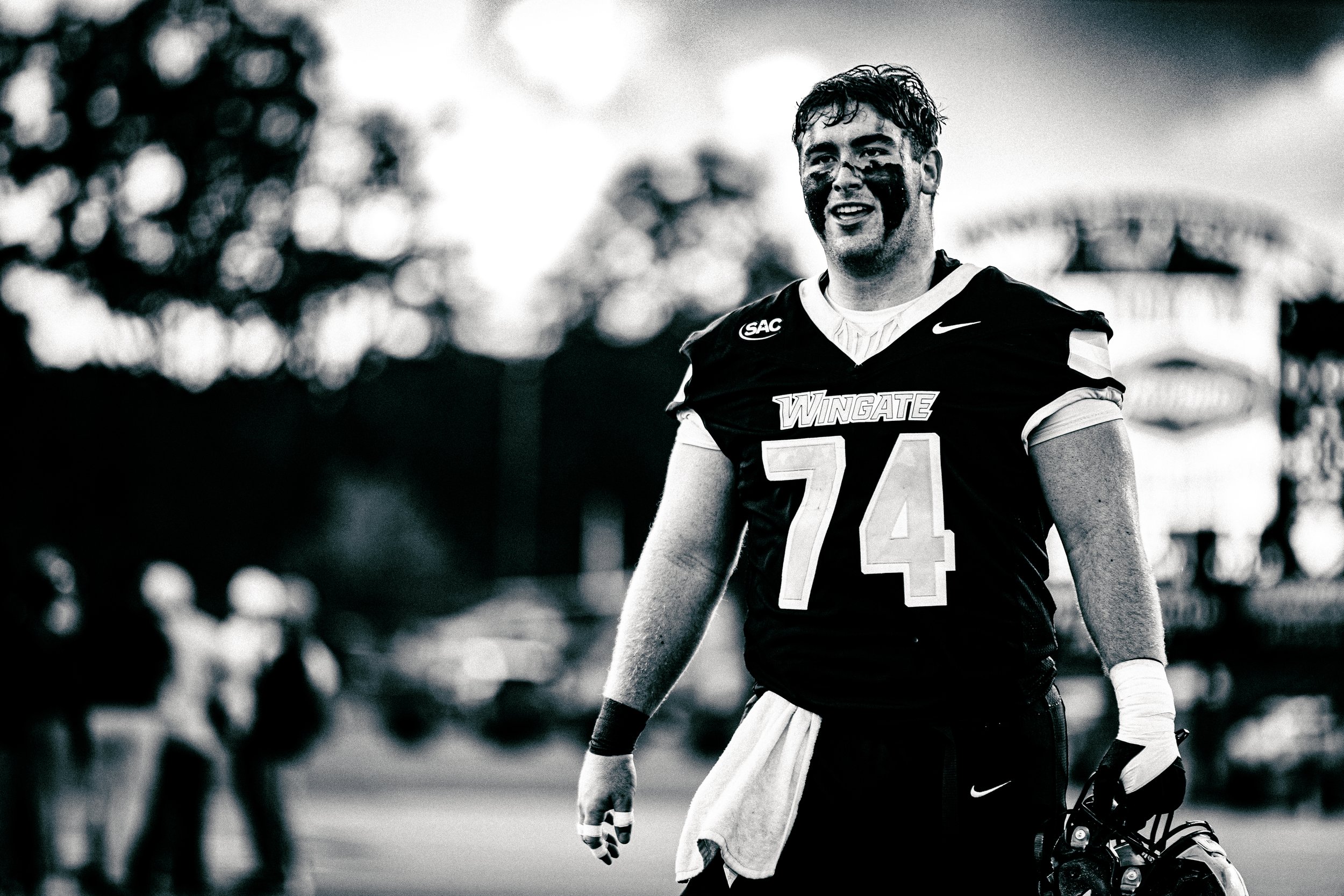 A football player stands outdoors with a towel over his waist and a helmet and gloves in his hand, smiling, with blurry spectators or players in the background. The photo is black and white.