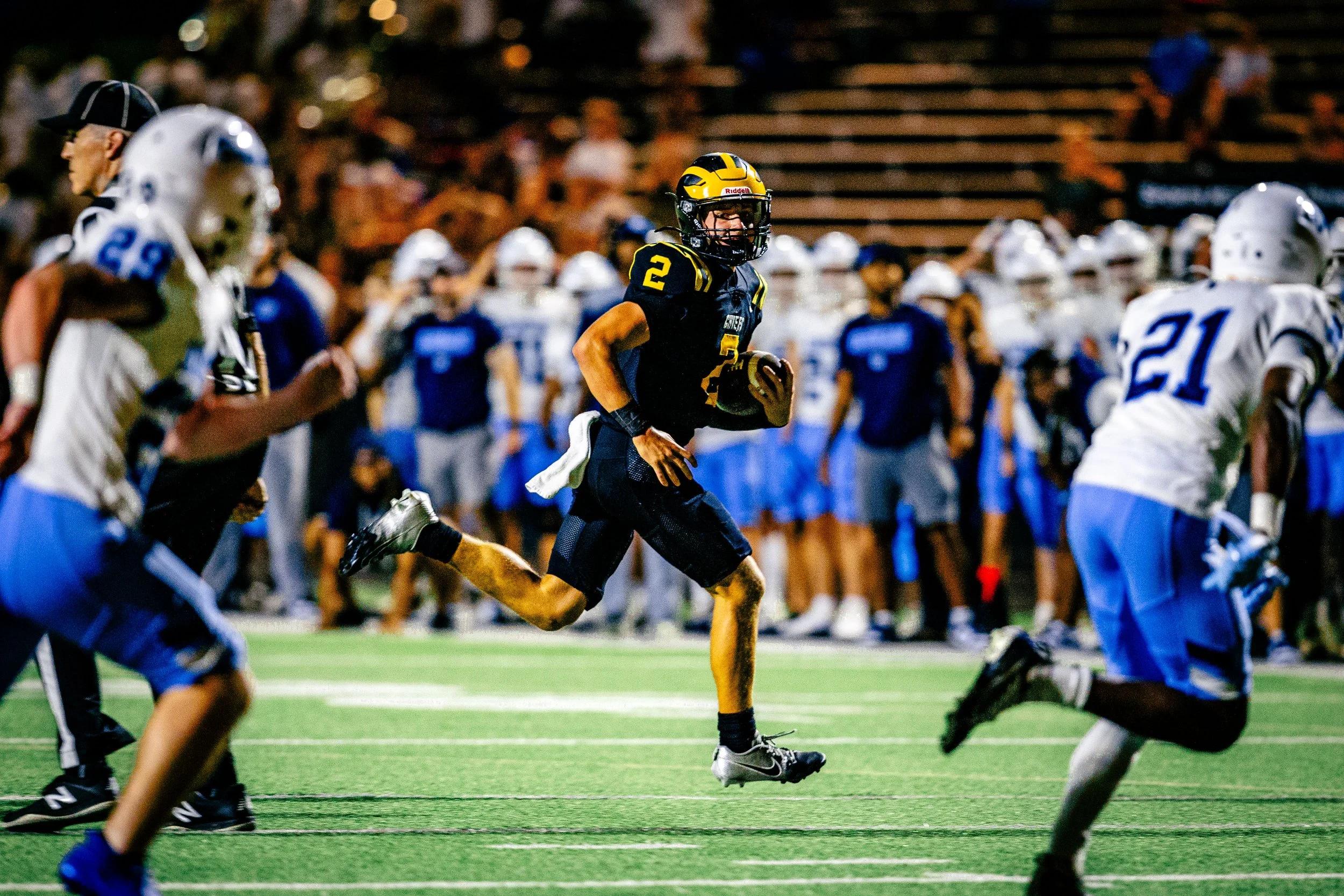 A football player wearing a black and yellow uniform runs with the ball, surrounded by players in white and blue uniforms, on a green field during a game at night.