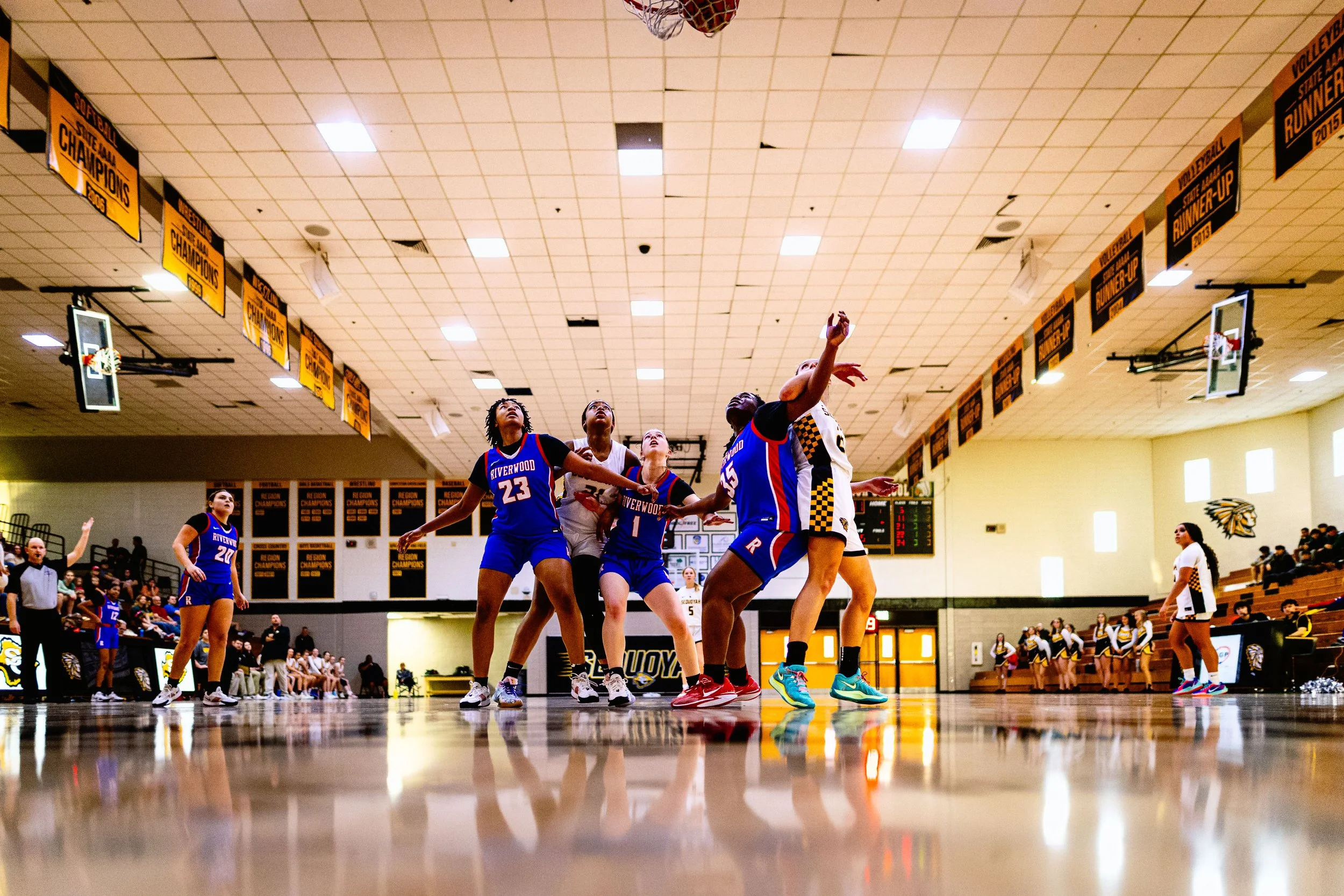 A girls' basketball game in progress inside a gymnasium, with players from two teams near the basket for a potential shot. The team in blue jerseys appears to be on offense, while the team in white and black uniforms is on defense. Spectators and che