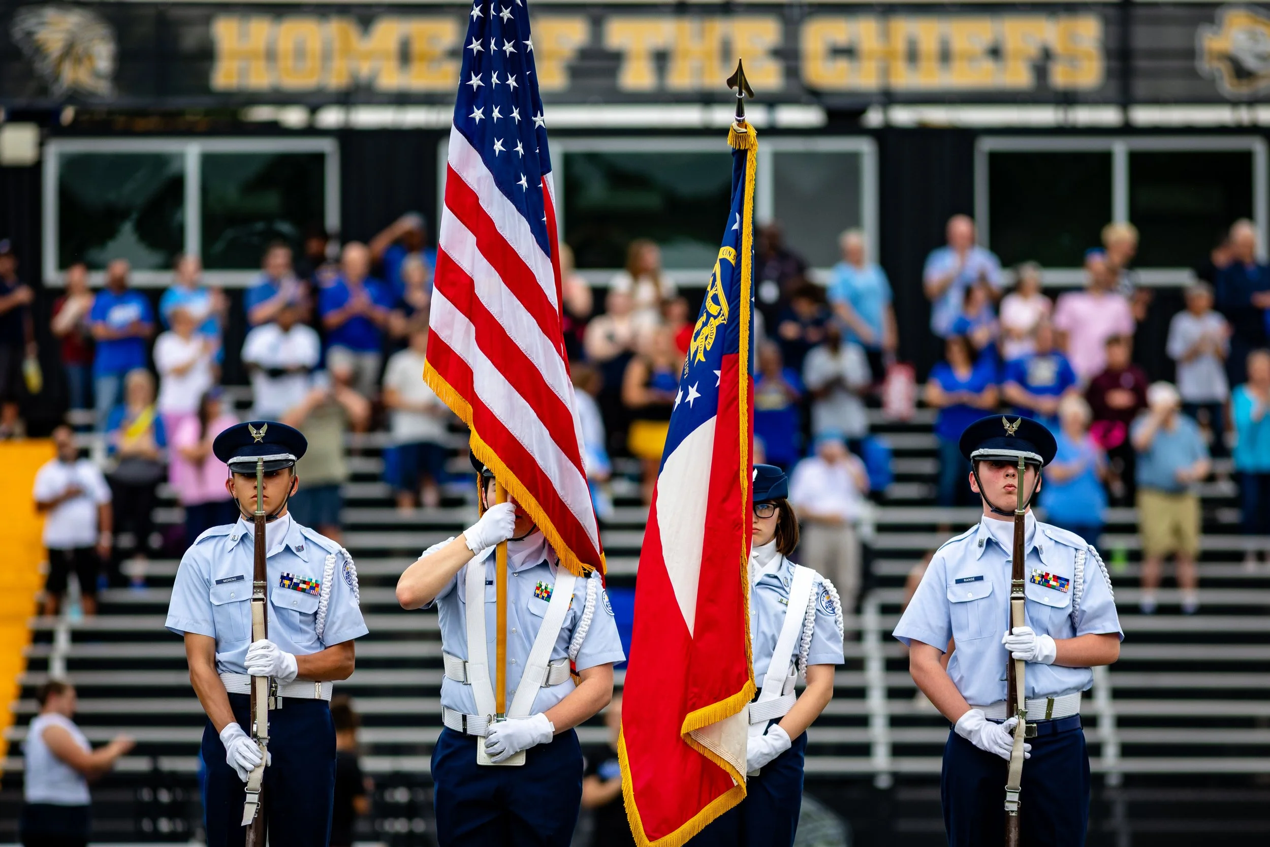 Four individuals in light blue military uniforms holding flags during a ceremony, with an audience in the background.
