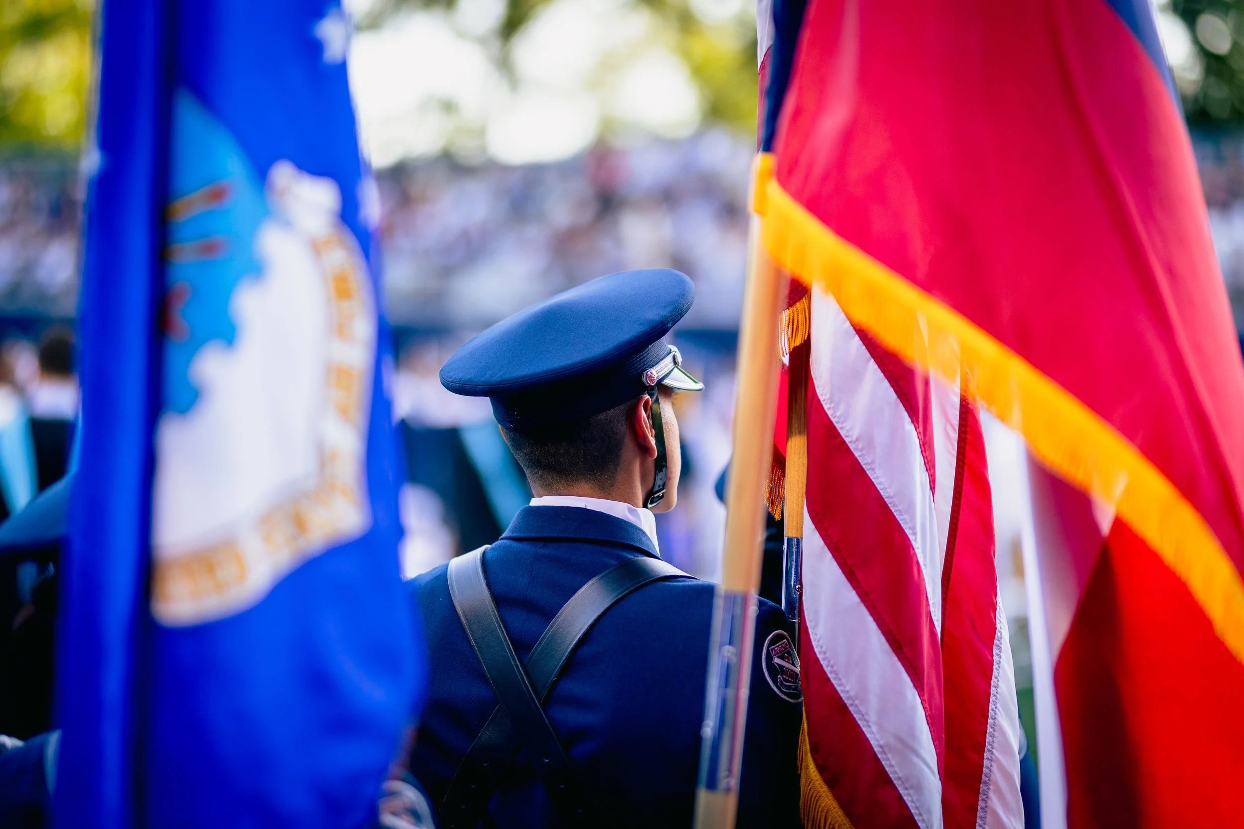 A person in a uniform holding a flag during a parade or ceremony, with American flags visible in the background.
