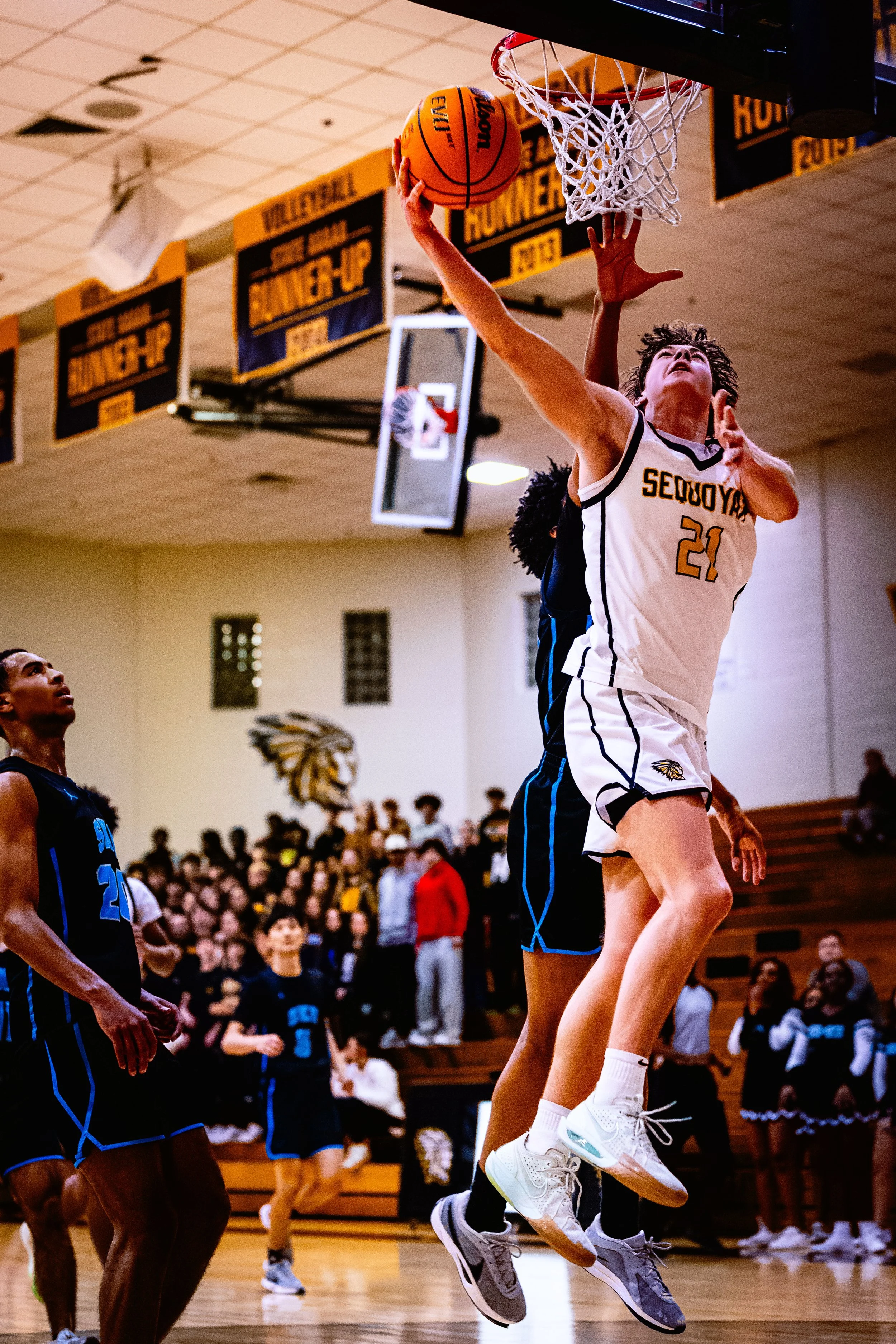 A basketball player in a white uniform with the number 21 is jumping to score a basket, with opponents in black and blue jerseys attempting to block the shot. Spectators watch from the bleachers.