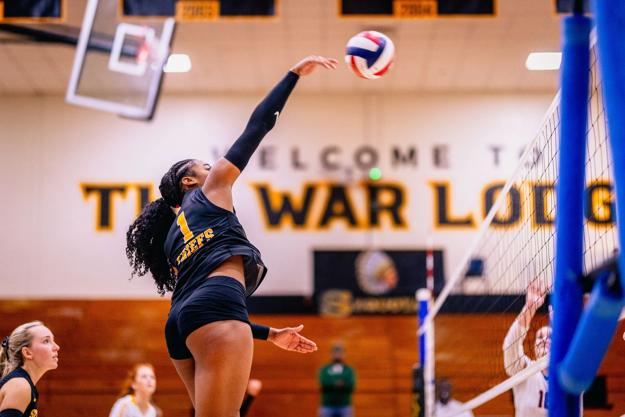 A female volleyball player in black uniform with the number 1 spikes the ball over the net during a game in a gymnasium.