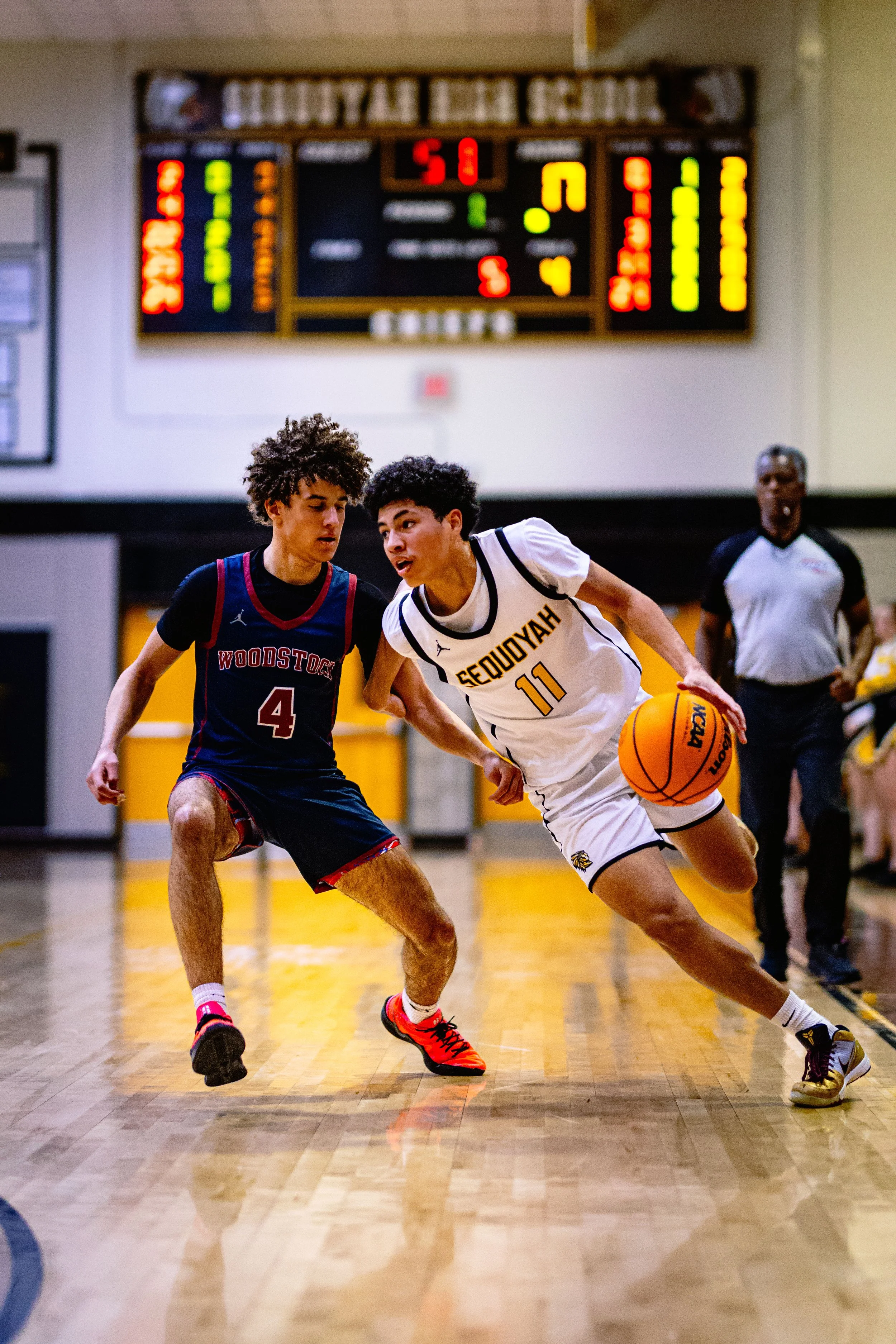 Two young male basketball players competing for the ball on a court with an electronic scoreboard in the background.