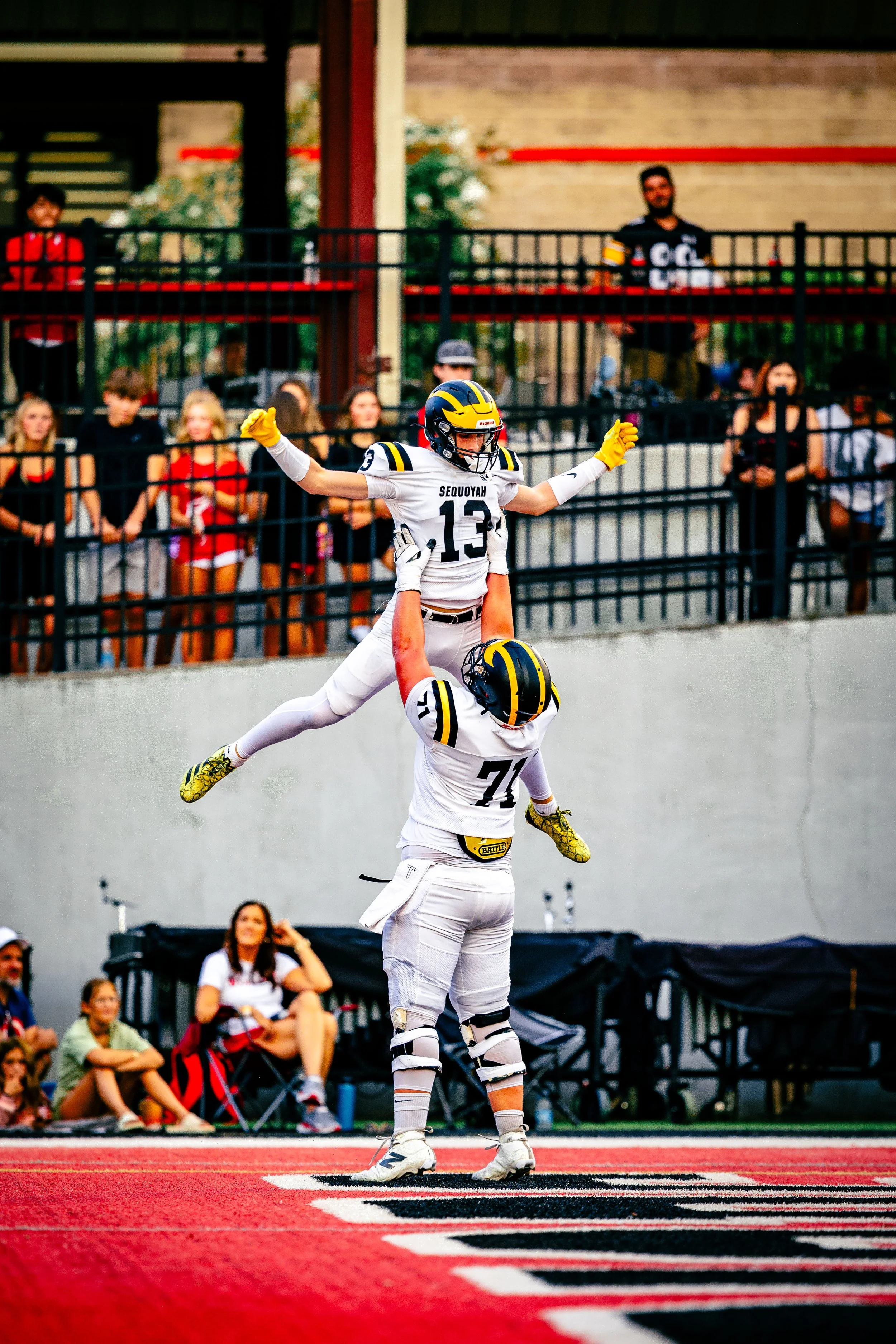 Two football players in white uniforms with black and yellow helmets celebrate on the field as one player lifts the other above his head. Spectators watch from the stands.