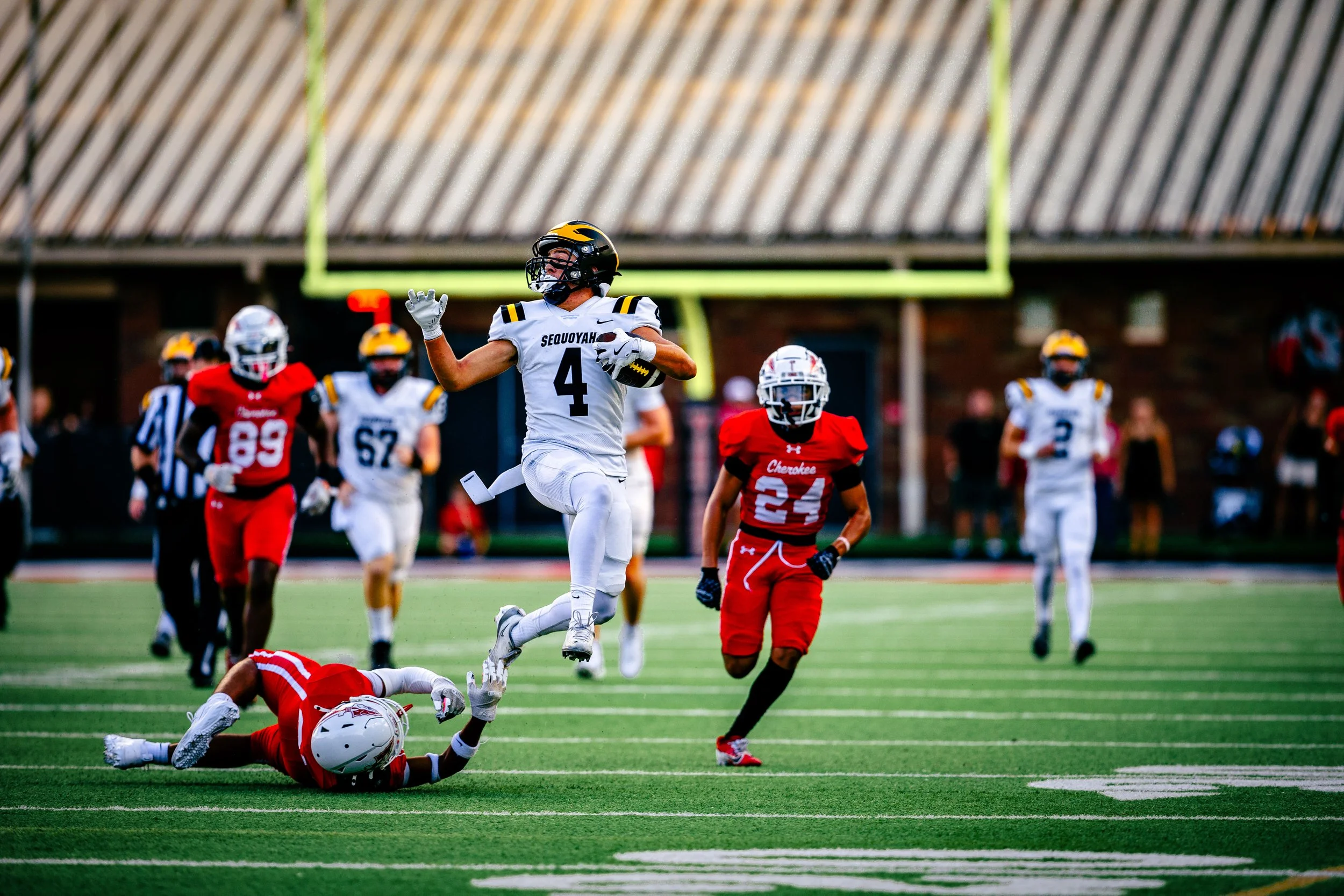 A football player wearing number 4 in a white jersey is running with the ball, evading another player who has fallen on the ground. Other players in red and white jerseys are visible in the background on a football field.