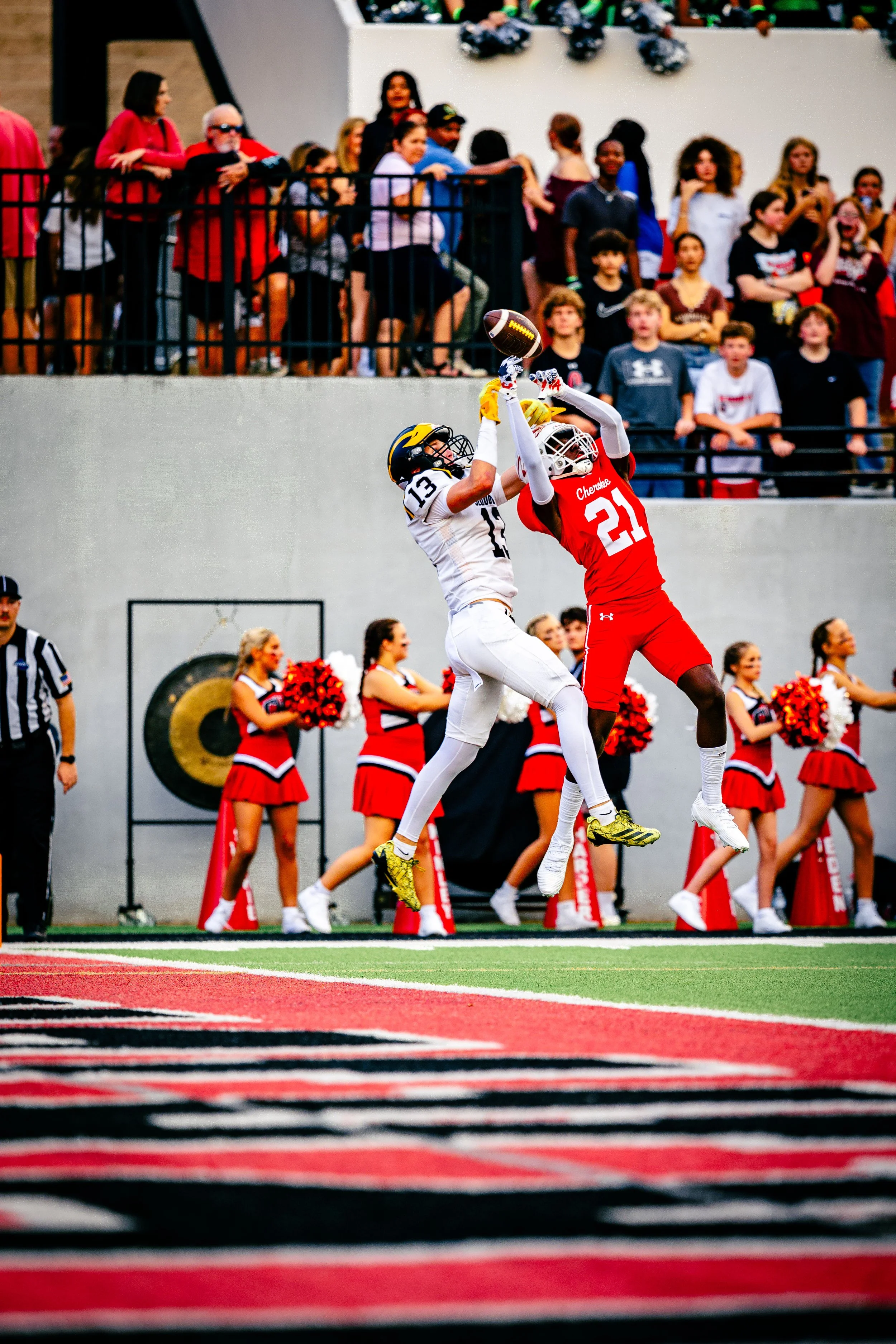 Two football players jump to catch the ball during a game, with cheerleaders and spectators watching in the background.