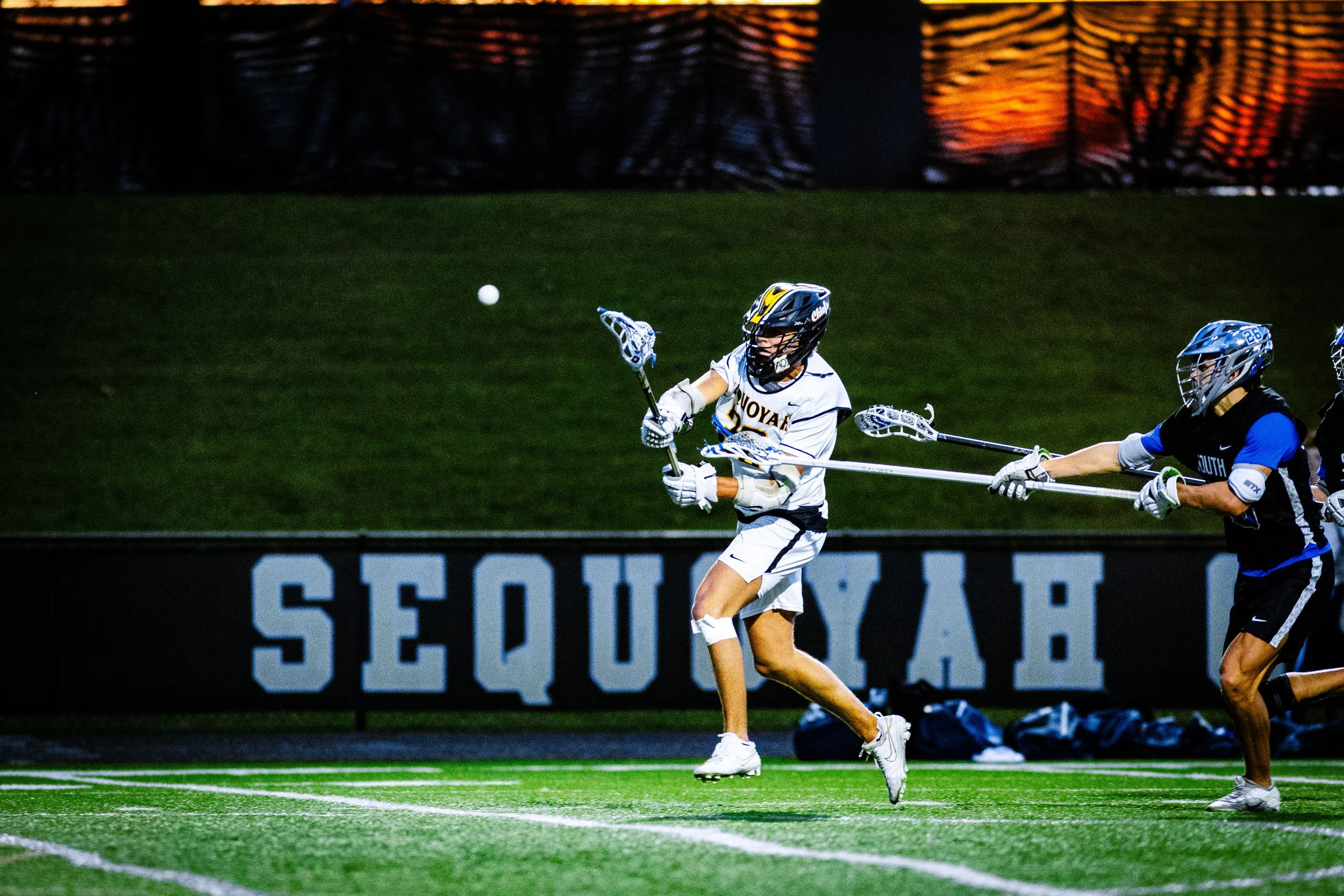 Lacrosse players in a game on a field during sunset, with one player preparing to catch a ball while another player reaches out.