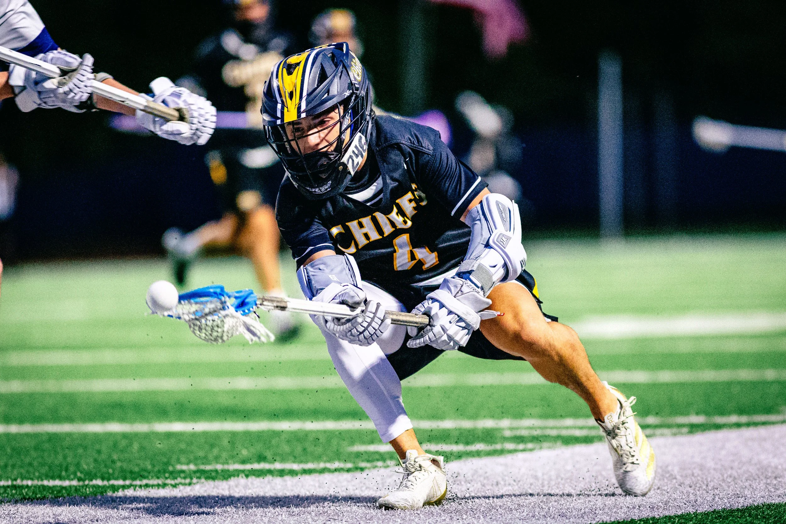 Lacrosse player in black uniform with yellow and white lettering and the number 4, wearing helmet and protective gear, lunging forward to reach the ball with the stick during a game at night.