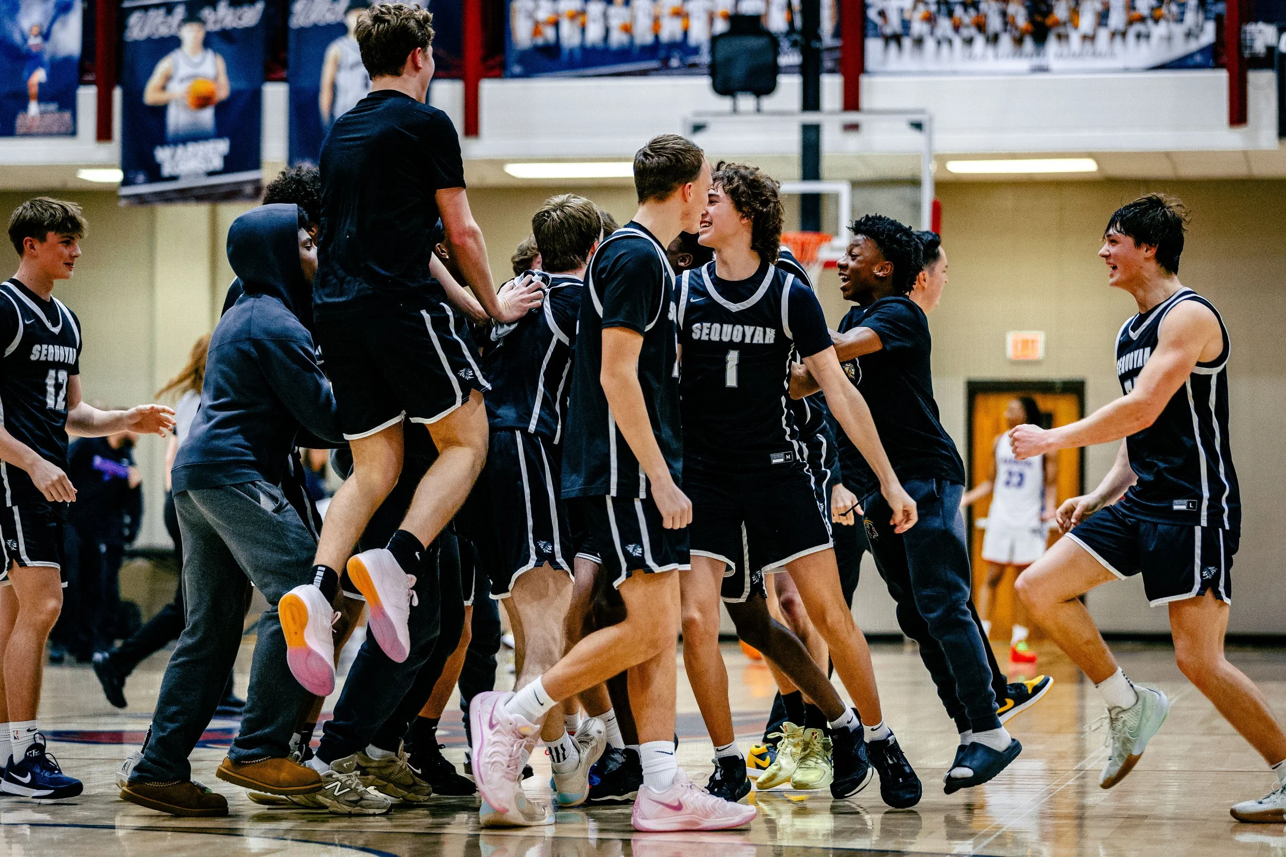 A group of young basketball players celebrating on the court, some jumping and others hugging, wearing black jerseys with white stripes and 'SEQUOYAH' written on them.