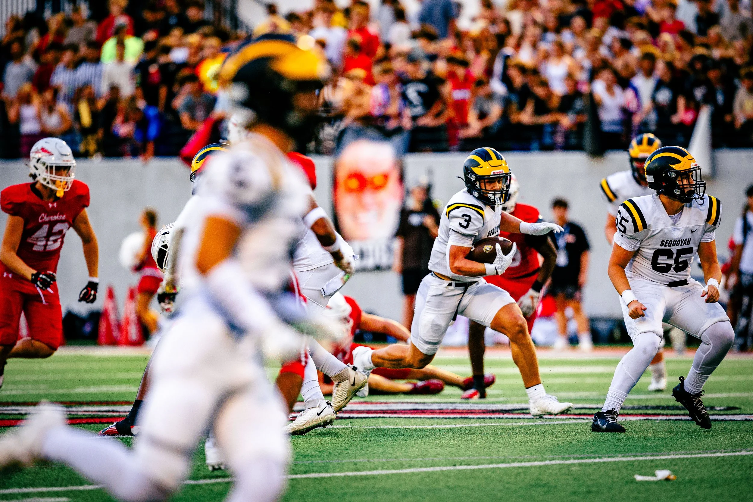 American football game with players running, including one carrying the ball, in a stadium with a large crowd in the background.