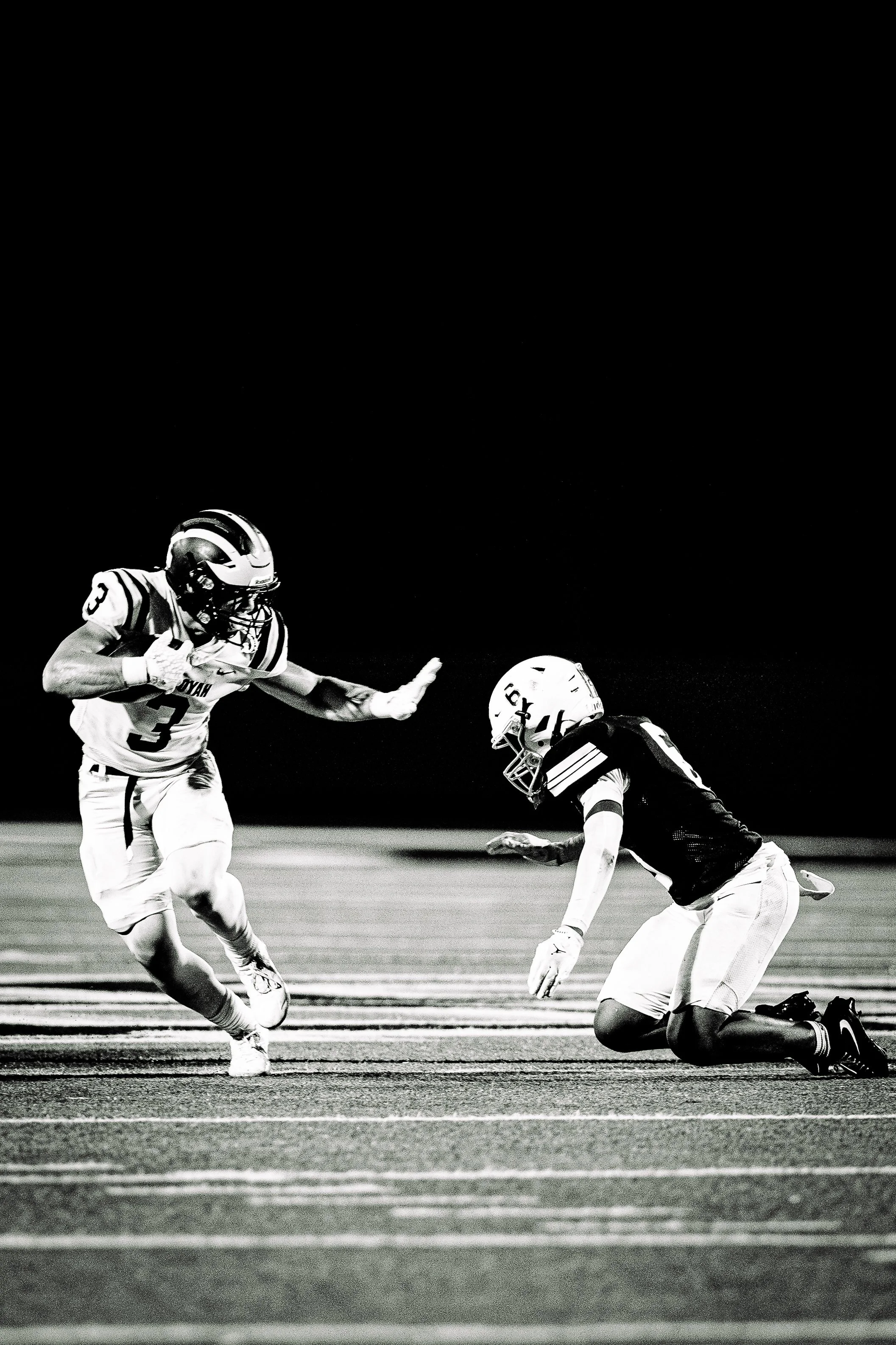 In this black-and-white image, a football game player wearing a jersey with the number 3 is running while a player from the opposing team, wearing a helmet and jersey, is kneeling on the ground. The running player extends his right arm, possibly to b