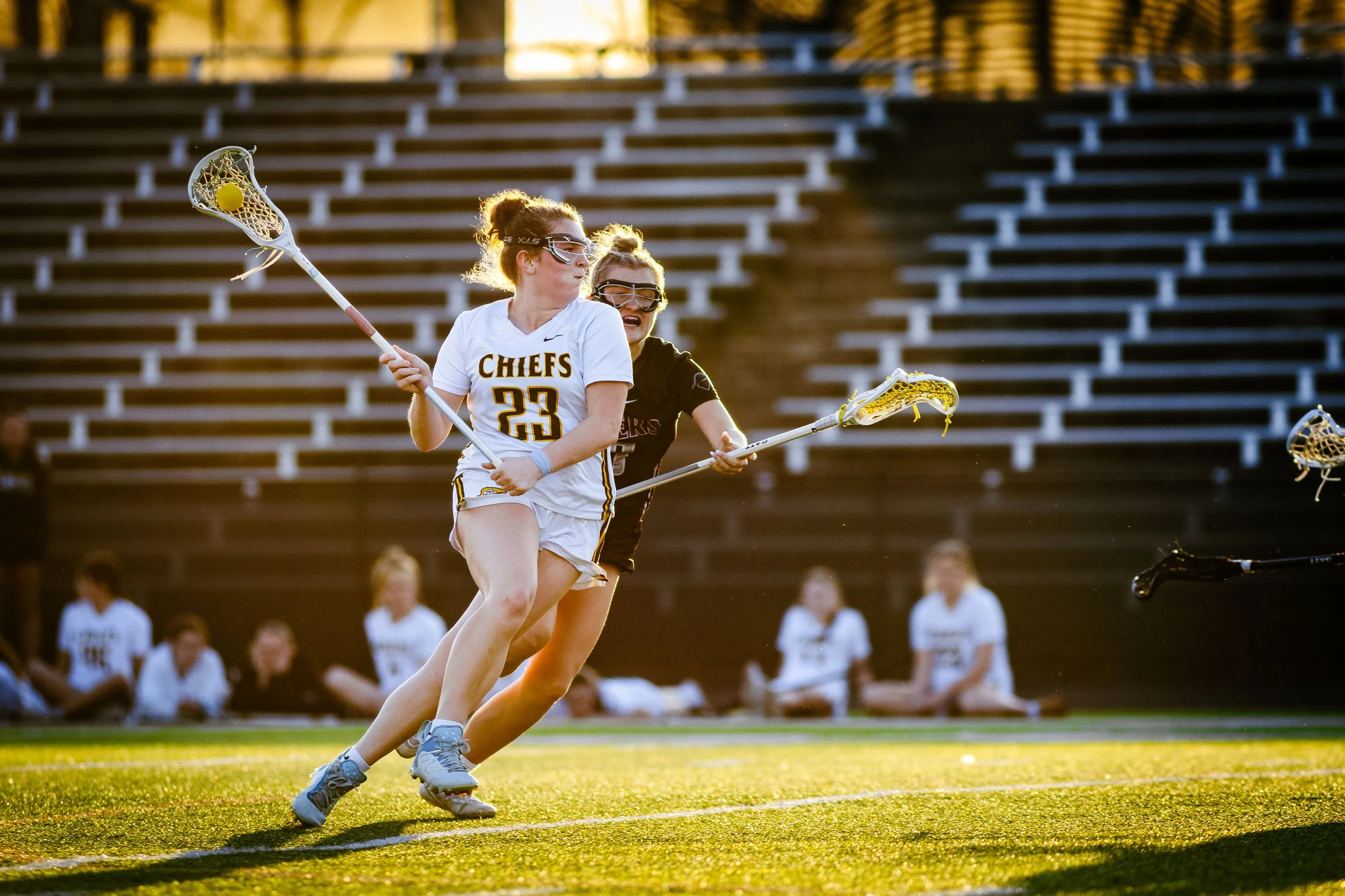 Two female lacrosse players in white and black uniforms compete for the ball during a game on a grass field, with empty bleachers in the background.