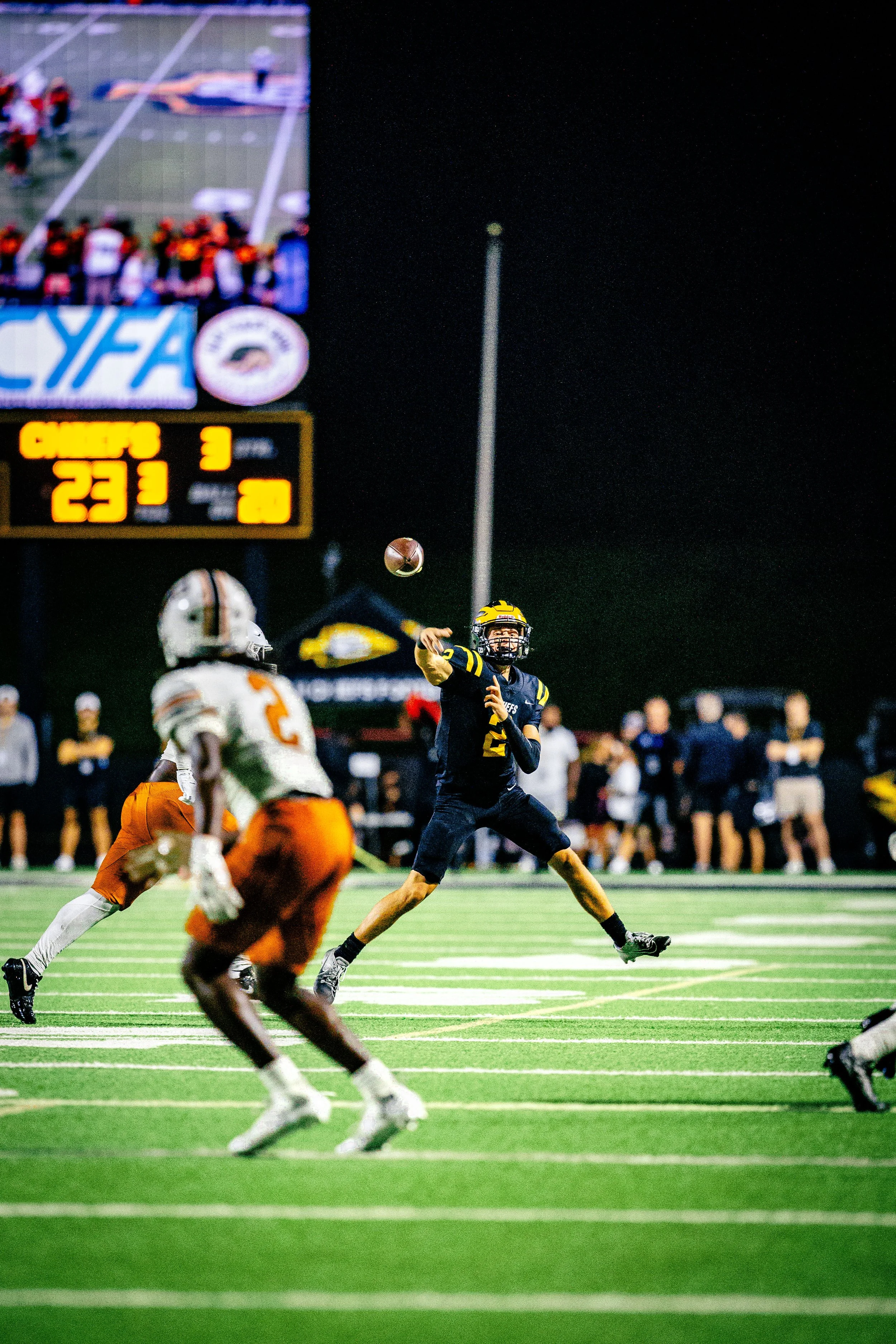 A football game at night, with a player in black uniform throwing the football and an opposing player in white and orange attempting to defend. The scoreboard in the background shows a score of 23-3 with 3 minutes remaining in the game.