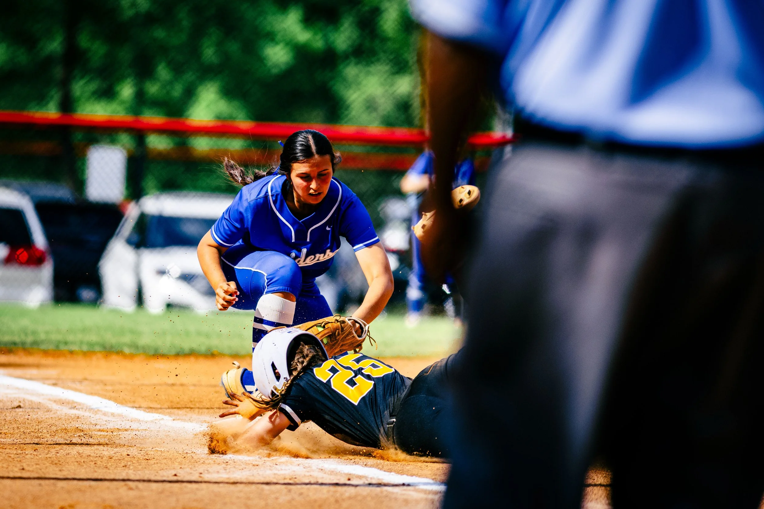 A female softball player in a blue uniform kneeling on the ground next to a sliding opponent in a dark uniform, with an umpire partially visible in the foreground, during a game on a dirt field.