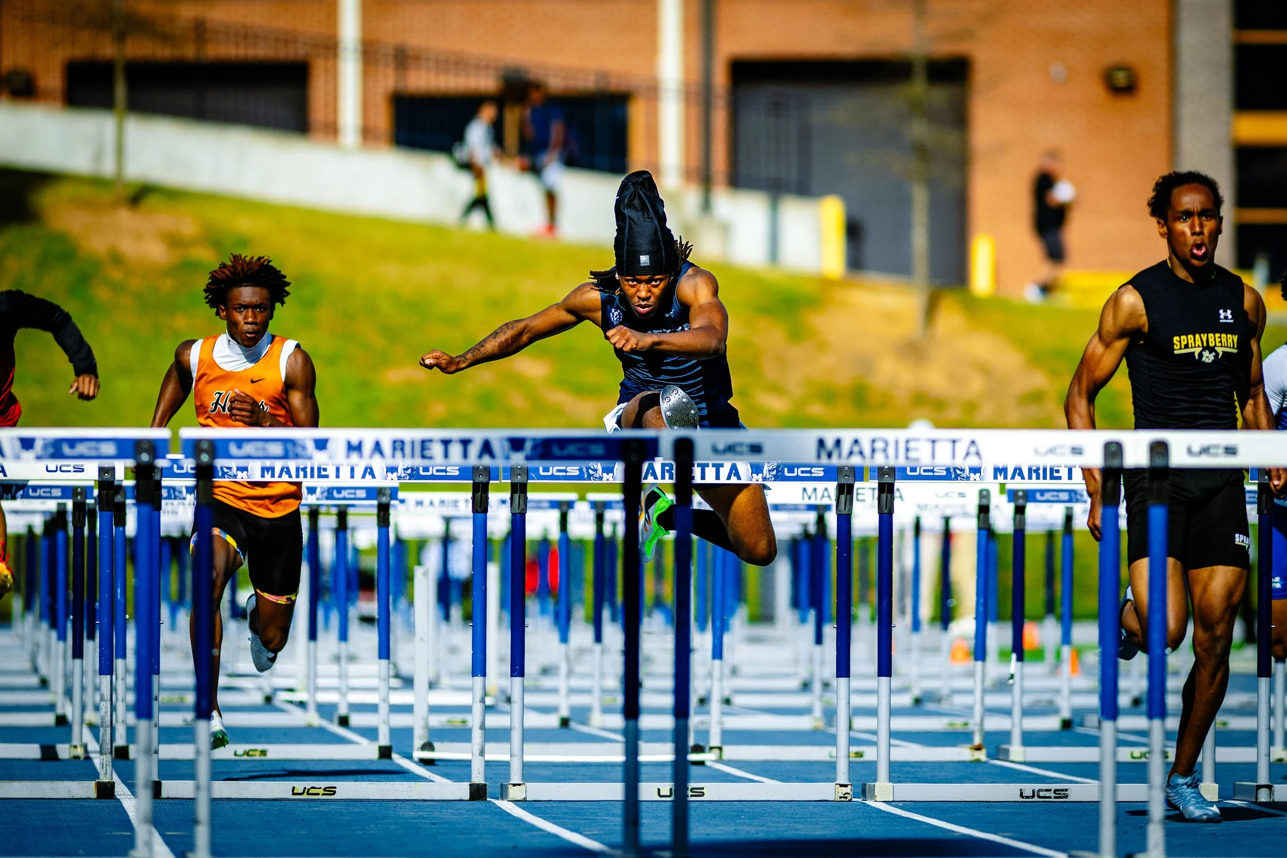 Track and field athletes competing in a hurdles race on an outdoor track, with a competitive focus and intense expressions, surrounded by grass and a building in the background.