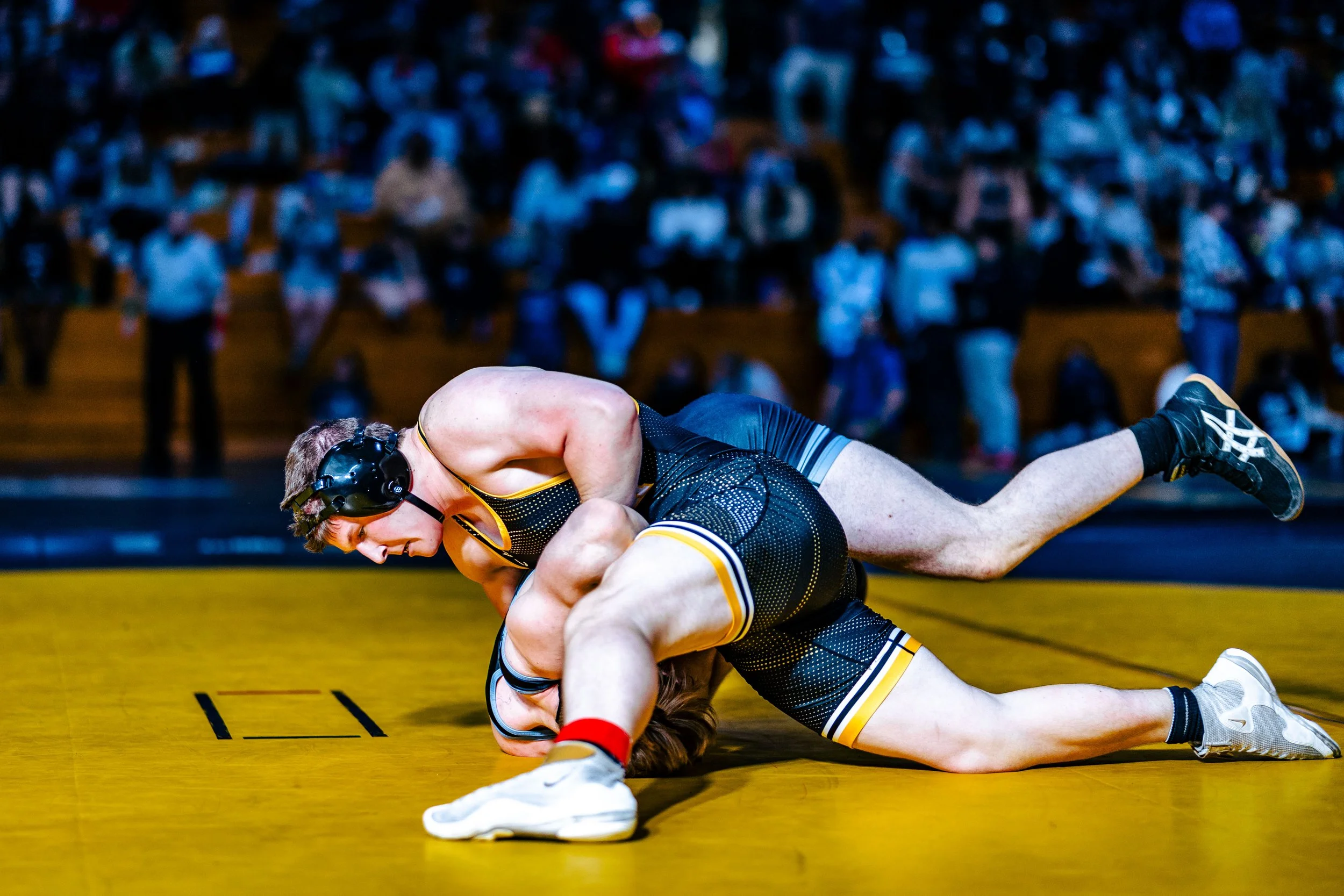 Two male wrestlers compete on a yellow wrestling mat in a gym, with a crowd watching in the background.